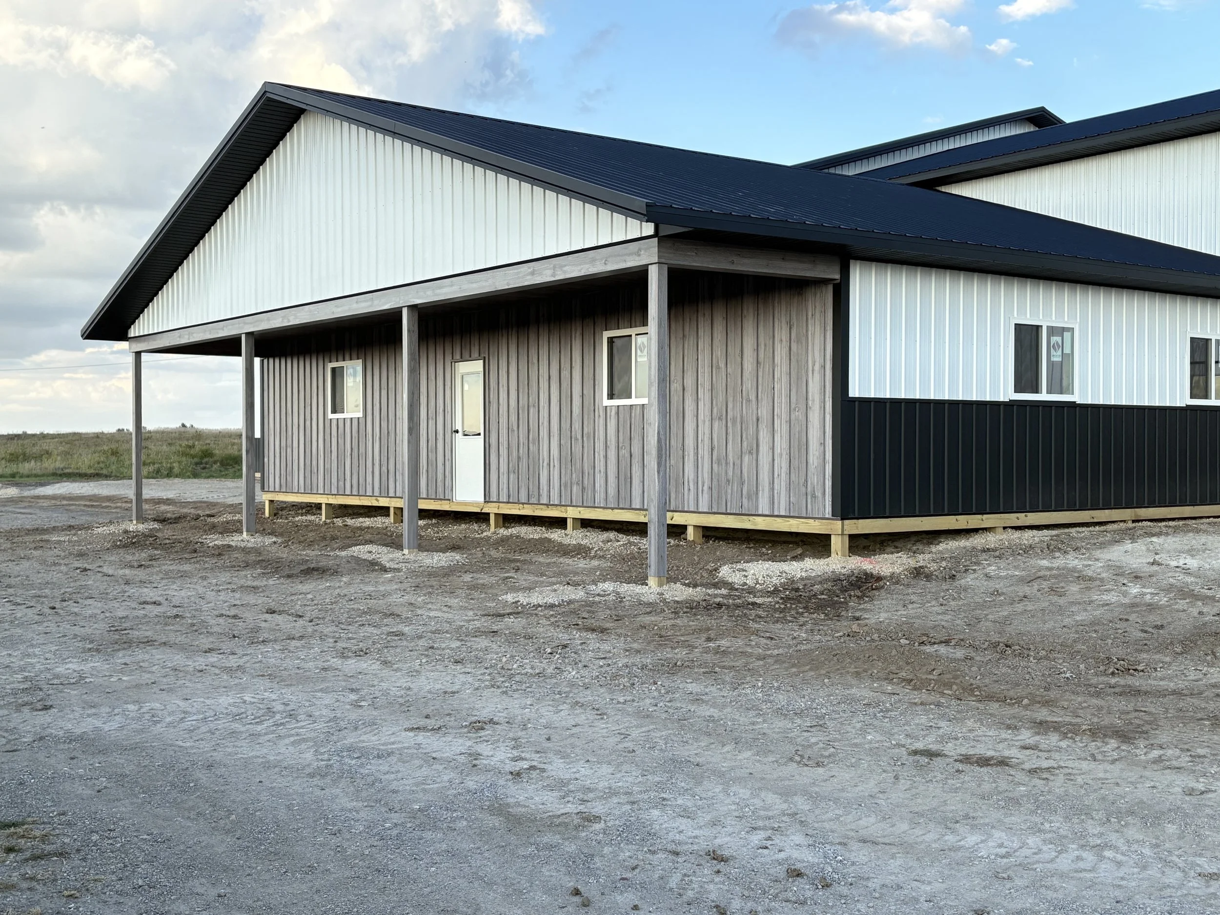 A modern house with a black metal roof and a combination of gray and white vertical siding on the exterior walls, located on a gravel lot under a partly cloudy sky.