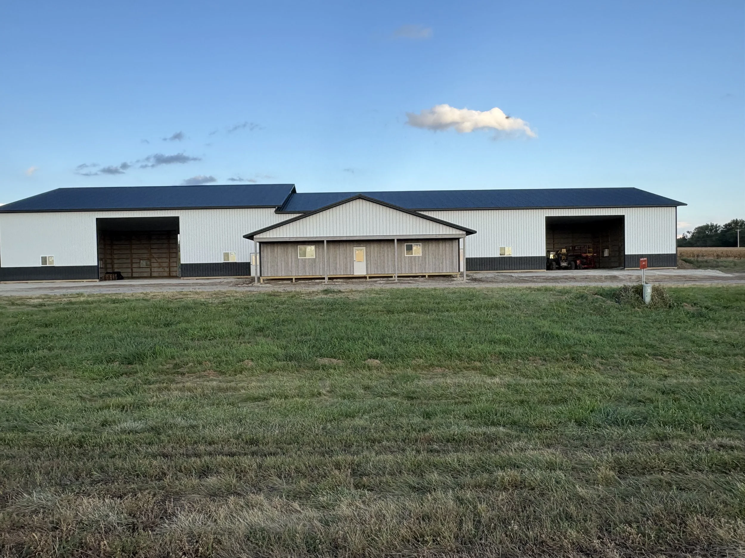 A large post frame barn building with two open garage doors, a small attached wooden structure, and a grassy foreground under a blue sky with scattered clouds.