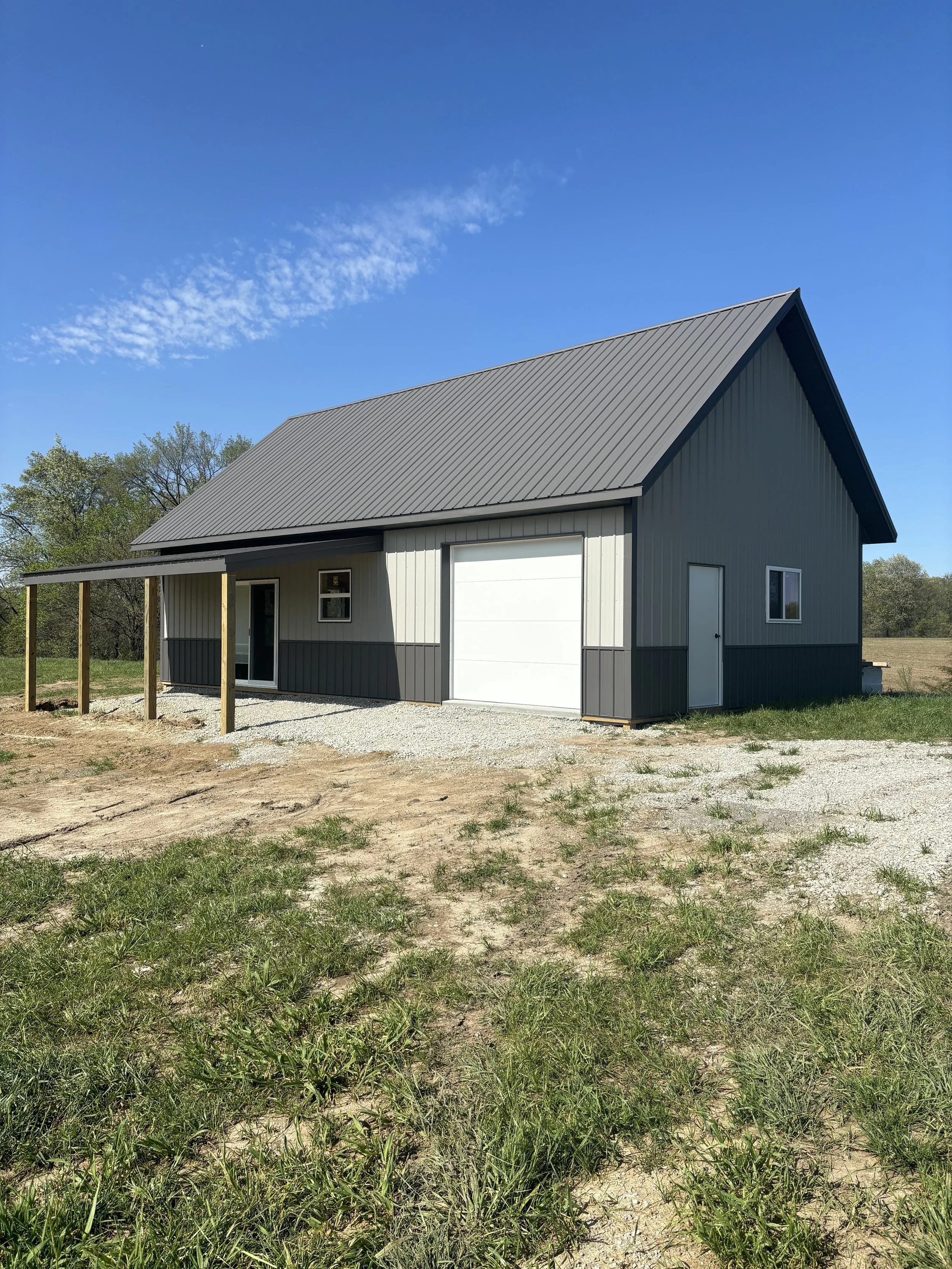A small metal building with a sloped roof and a white garage door, surrounded by grass and trees under a clear blue sky.