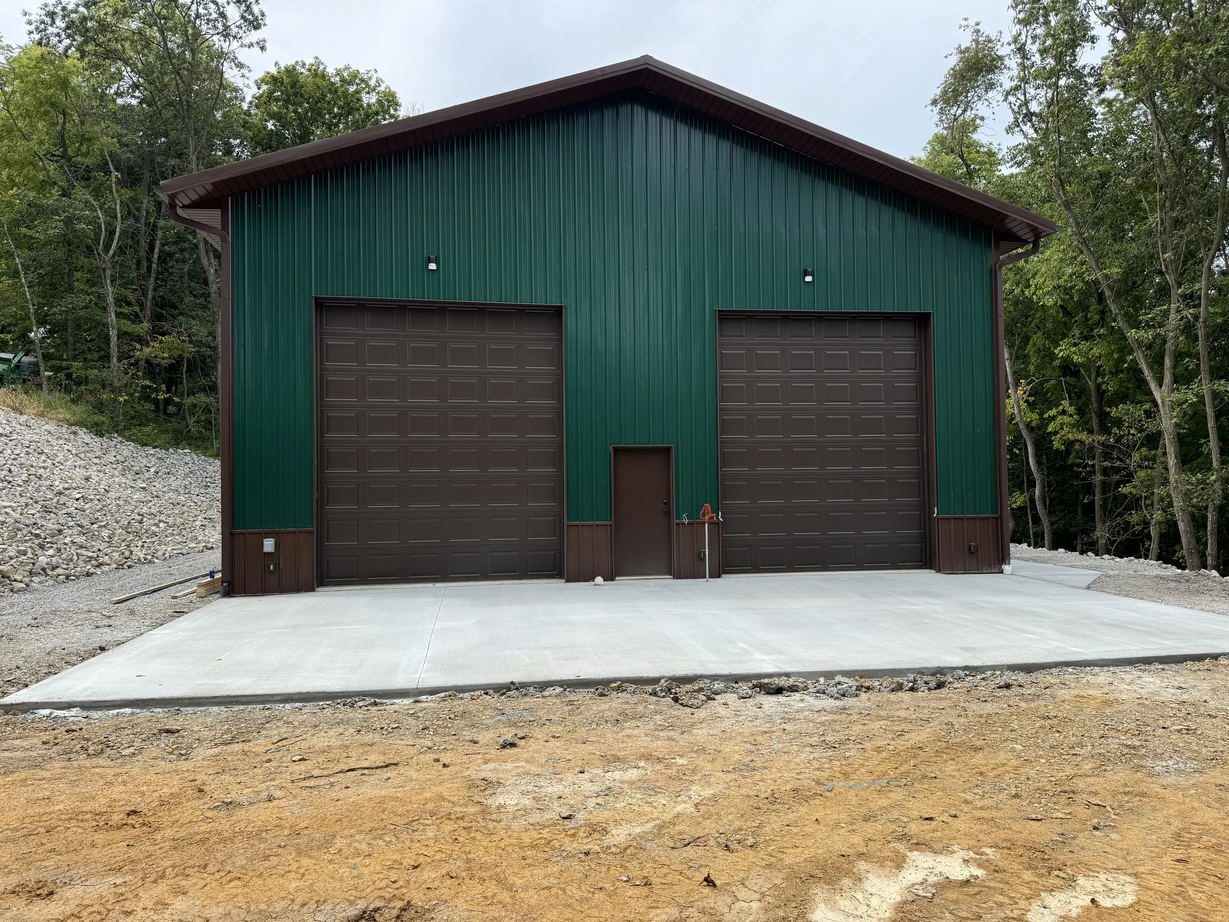 Large green and brown metal building with two garage doors, set against a background of trees and an overcast sky, with a freshly poured concrete driveway in the foreground.