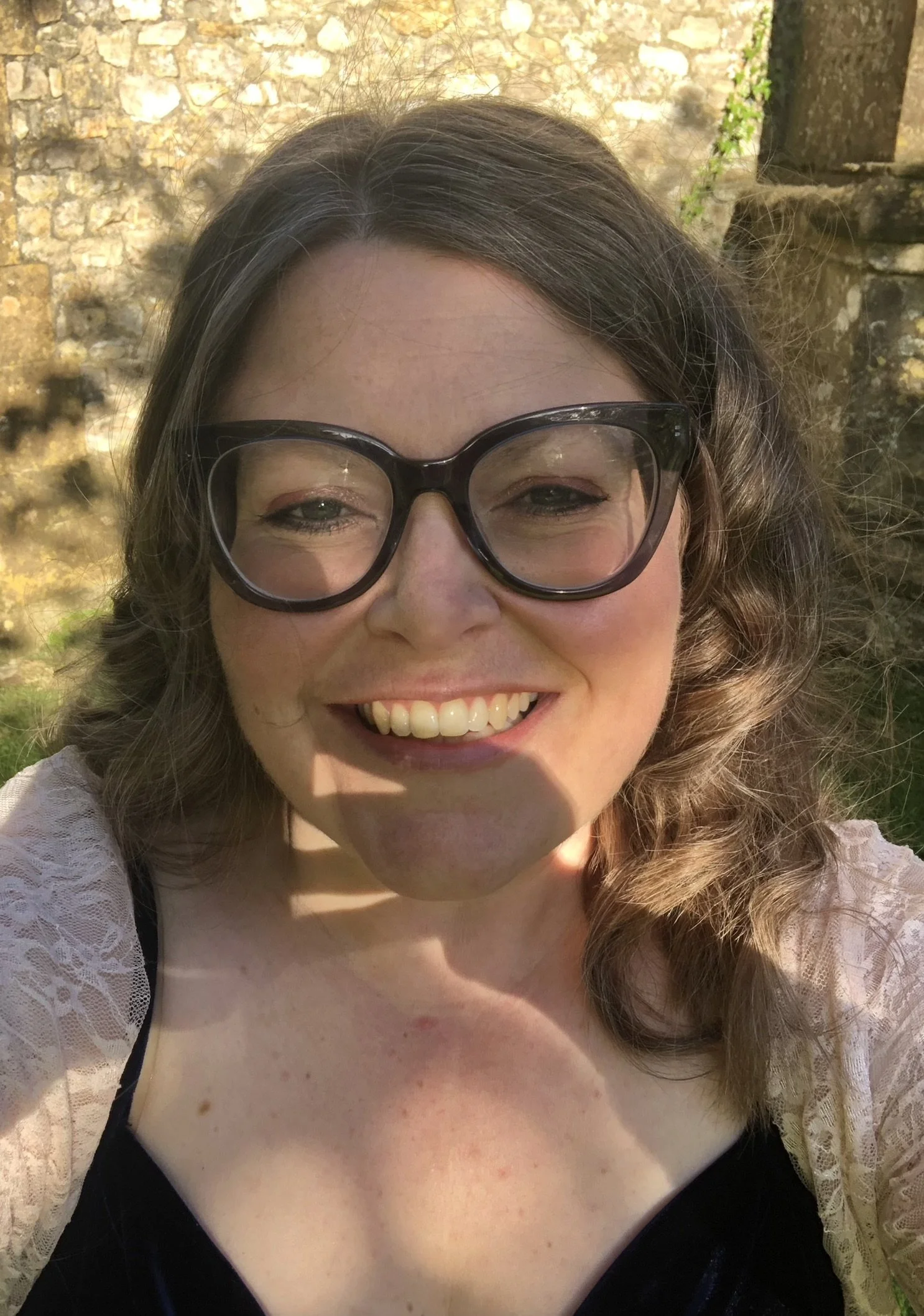 A woman with curly, brown hair and glasses smiling, outdoors in natural light, with a stone wall and some greenery in the background.