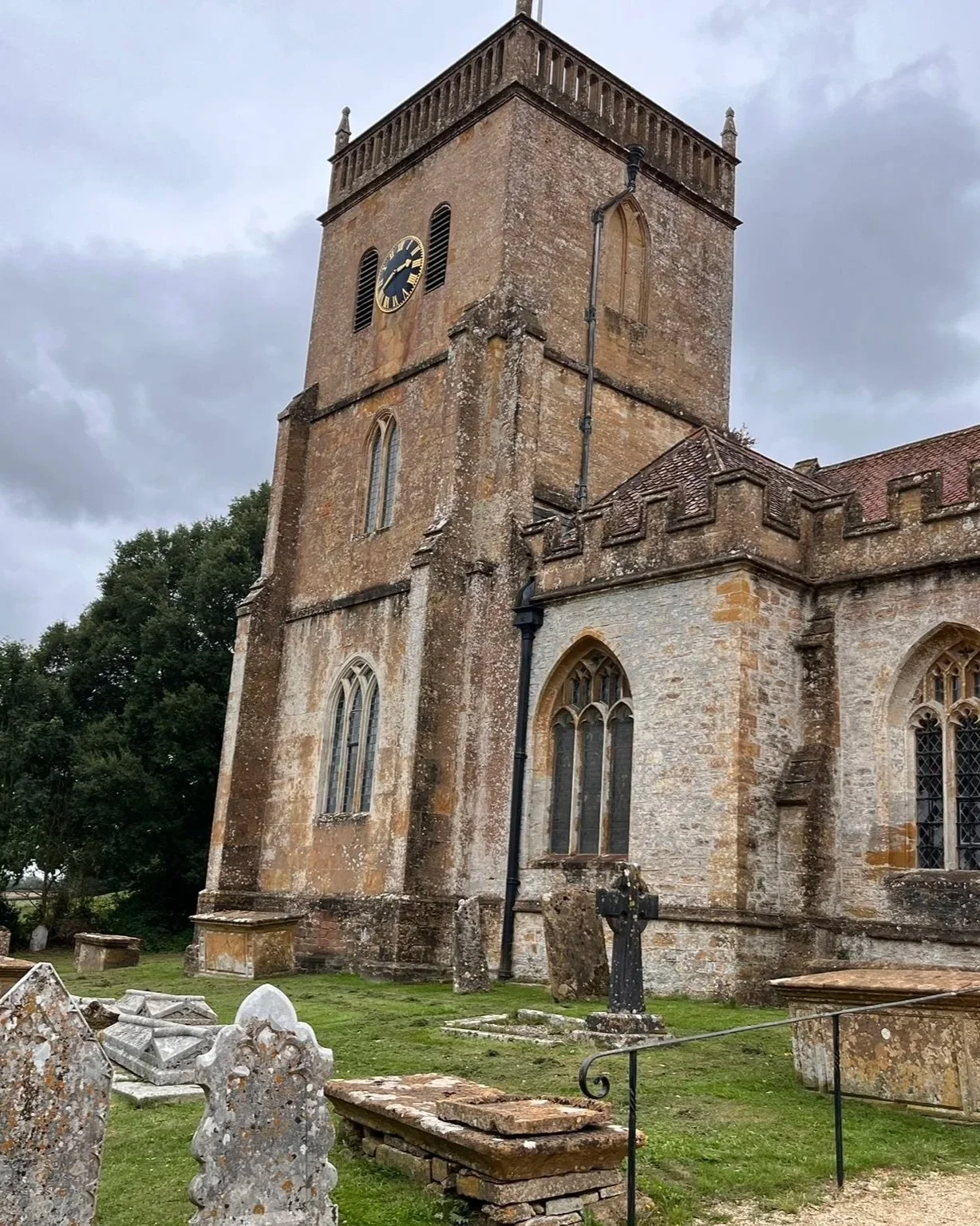 An old stone church with a tall clock tower, stained glass windows, and gravestones in the churchyard, under a cloudy sky.