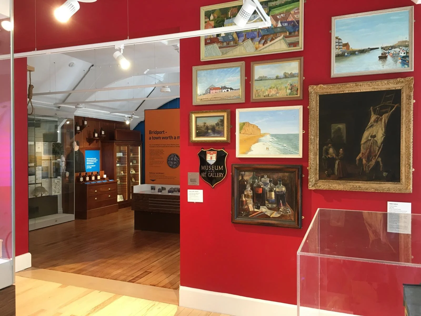 An art gallery with paintings on a red wall, including landscapes, a harbor scene, and a dark painting of a horse, with gallery signage and display cases in the background.
