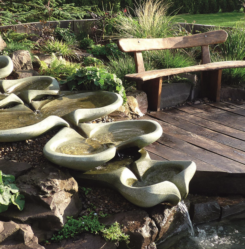 A garden scene with a tiered stone water fountain, a wooden bench, and lush greenery.