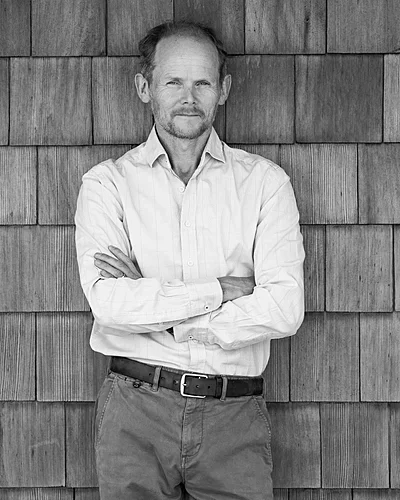 A man with a beard and mustache wearing a dress shirt with sleeves rolled up and dark pants, standing with arms crossed in front of a wooden wall.