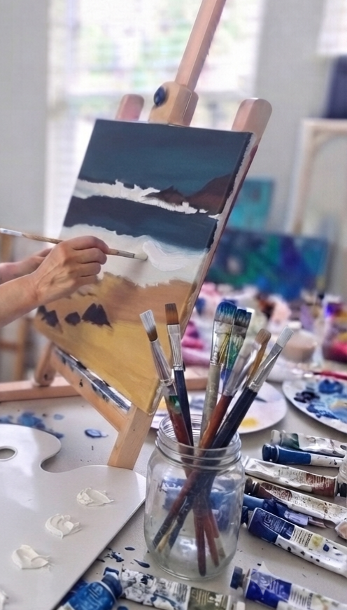 Person painting a landscape with mountains and water on an easel in an art studio.