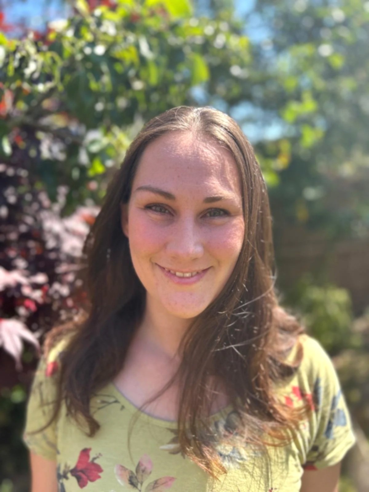 Smiling woman with long brown hair standing outdoors on a sunny day, with green trees and pink flowers in the background.