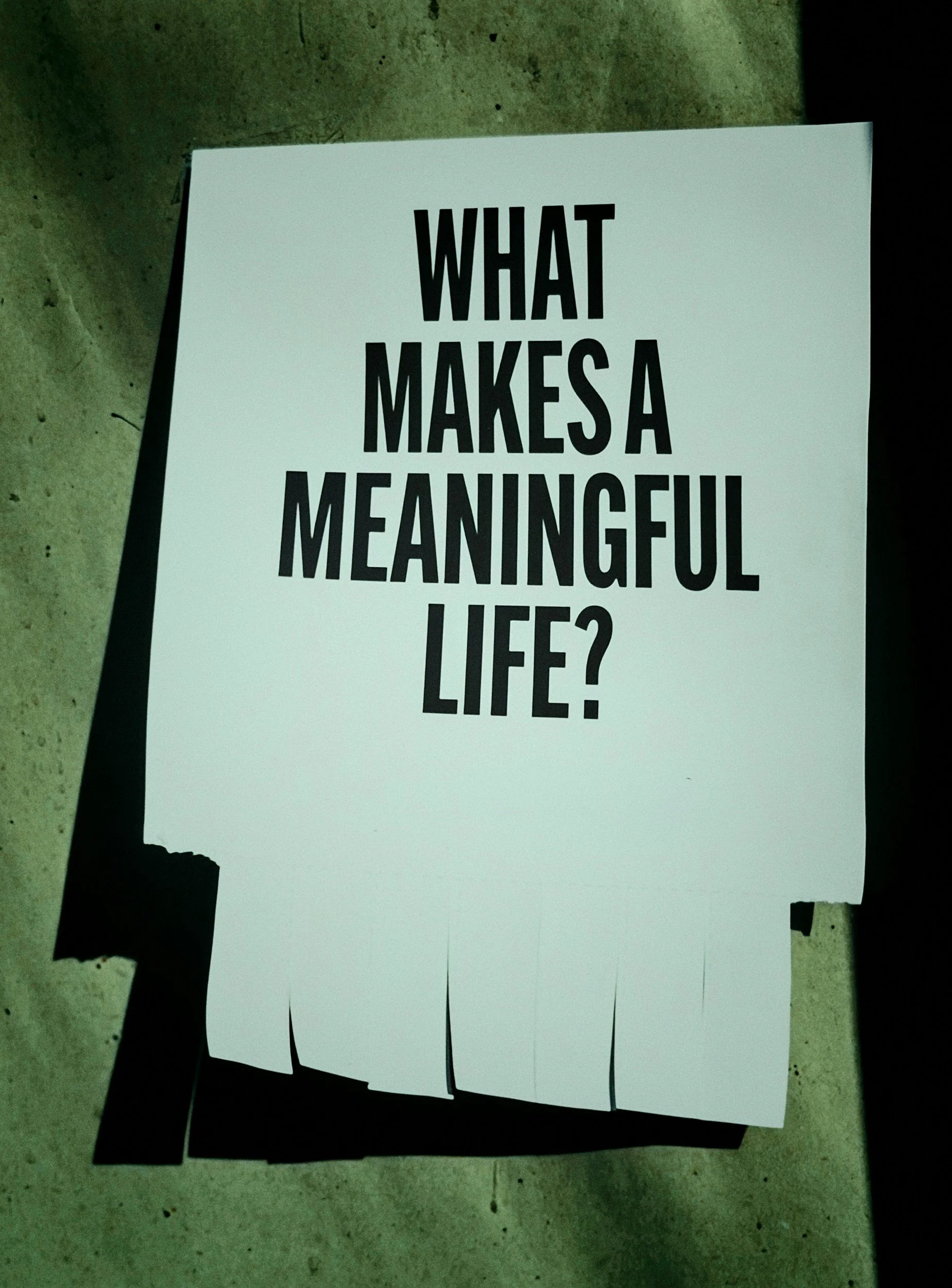 A stack of white paper sheets with the bold black text: "What makes a meaningful life?" printed on the top sheet. The sheets are placed on a concrete floor.