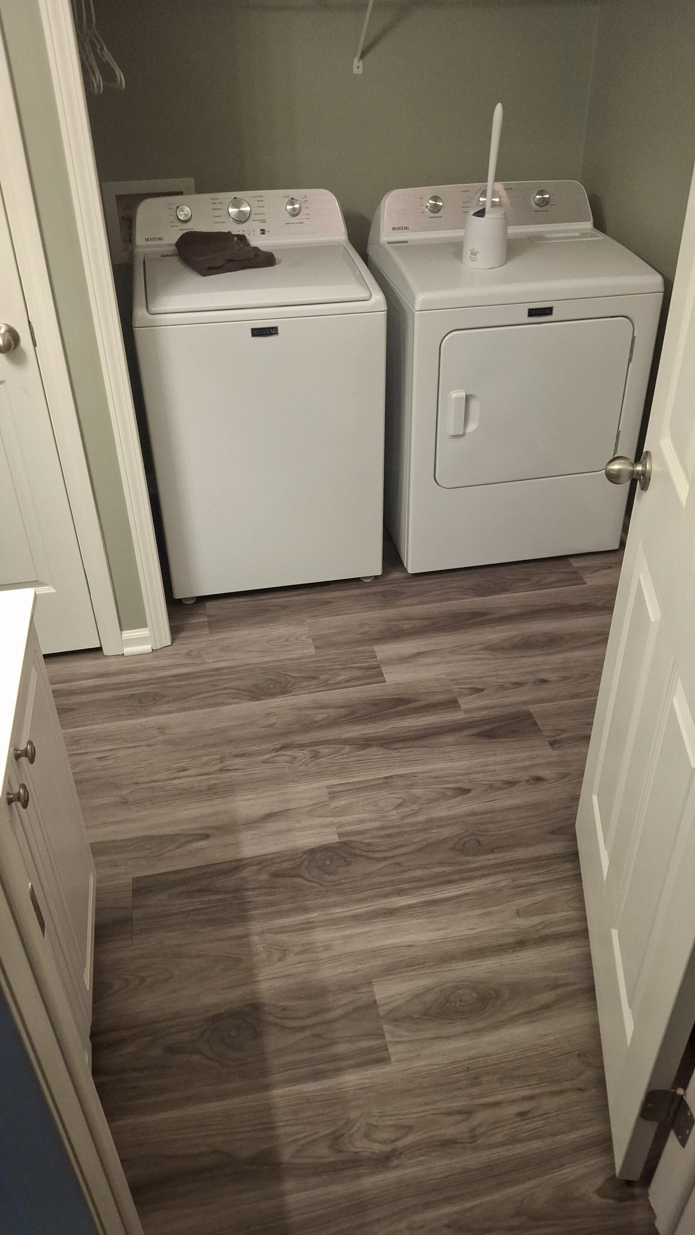 A laundry room with a white washing machine and a white dryer next to each other on a wooden floor. There is a brown cloth on top of the washing machine and a roll of paper towels in a white container on top of the dryer.