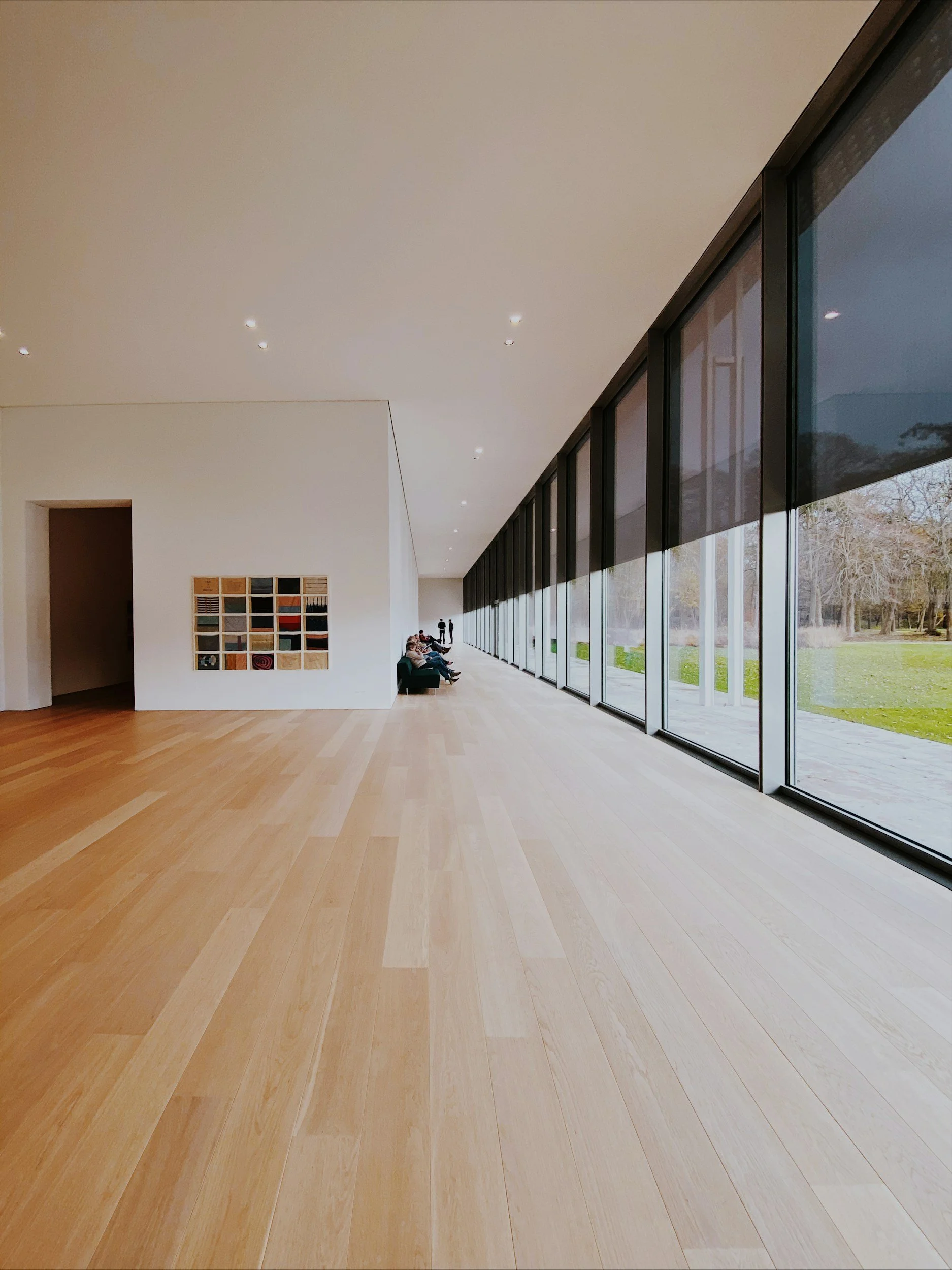 Interior of a modern building with wood flooring, white walls, large glass windows, and a few people sitting and walking.