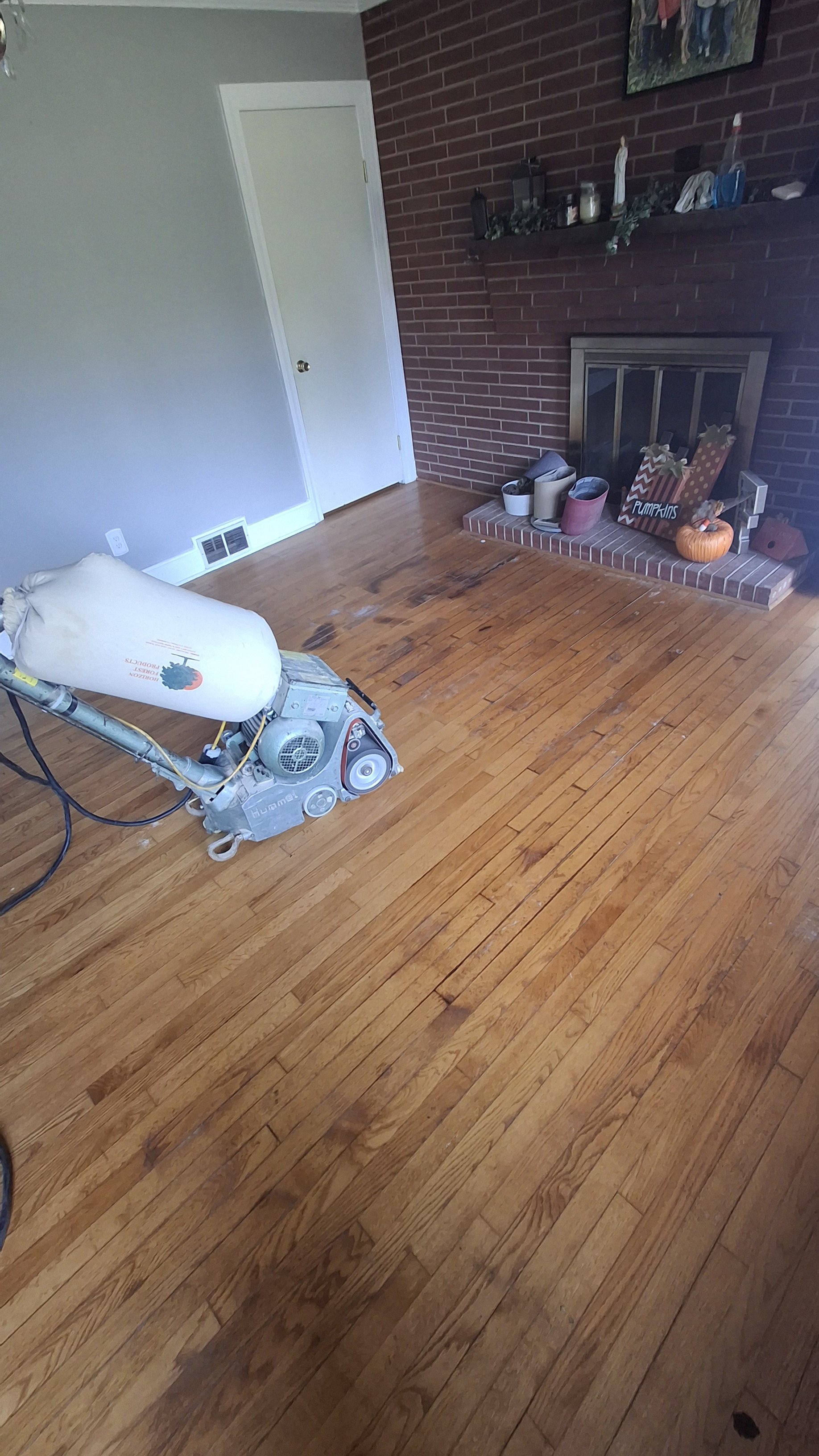 A room with wooden flooring, a brick fireplace decorated with pumpkins, and a cleaning machine in the foreground.