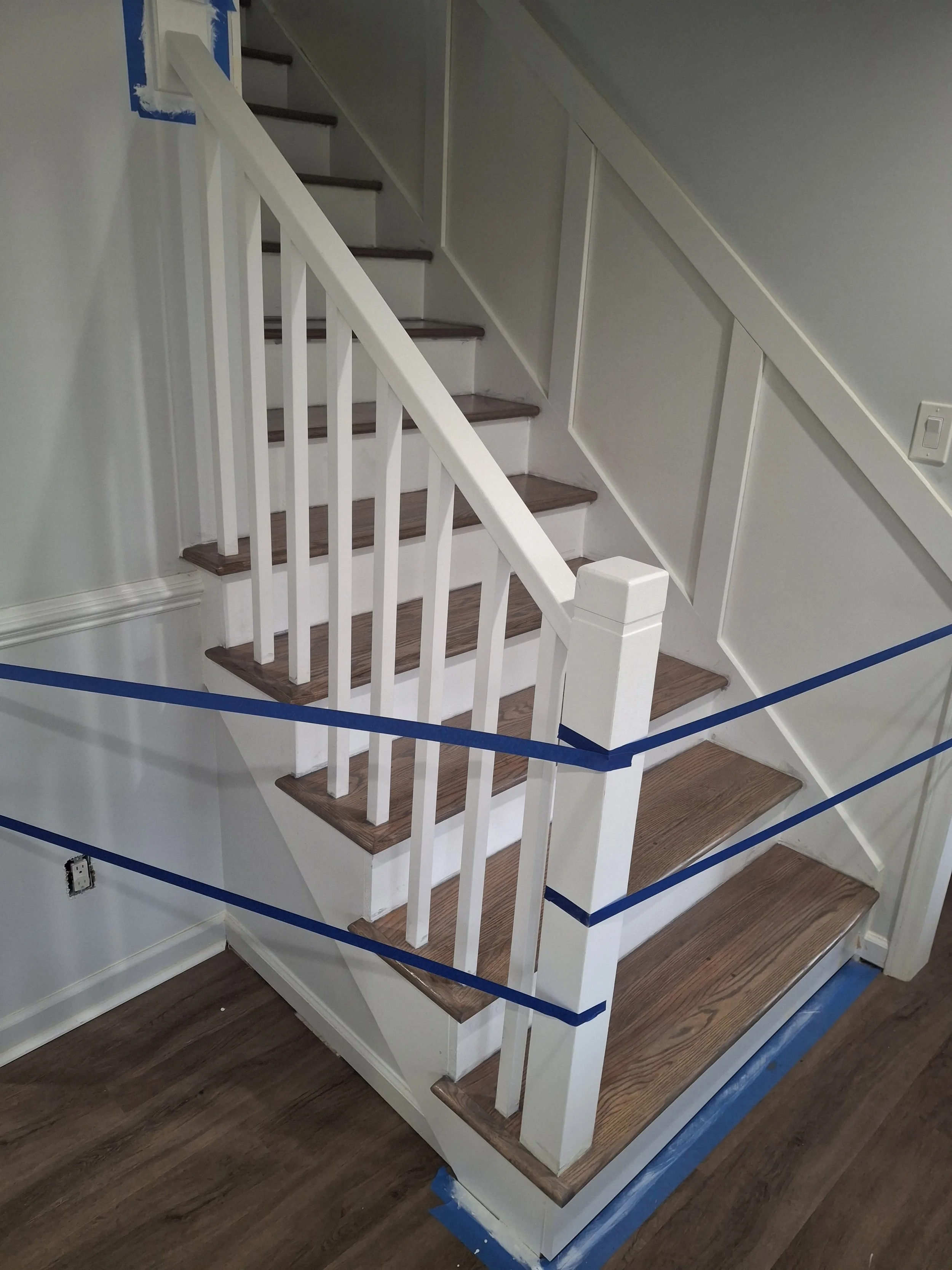 A staircase with dark wooden treads, white risers, and a white railing with vertical balusters. The wall beside the staircase is painted light gray, and there are blue painter's tape strips indicating ongoing painting work. The floor is made of dark wood, and a section of the blue painter's tape is visible at the bottom of the image.