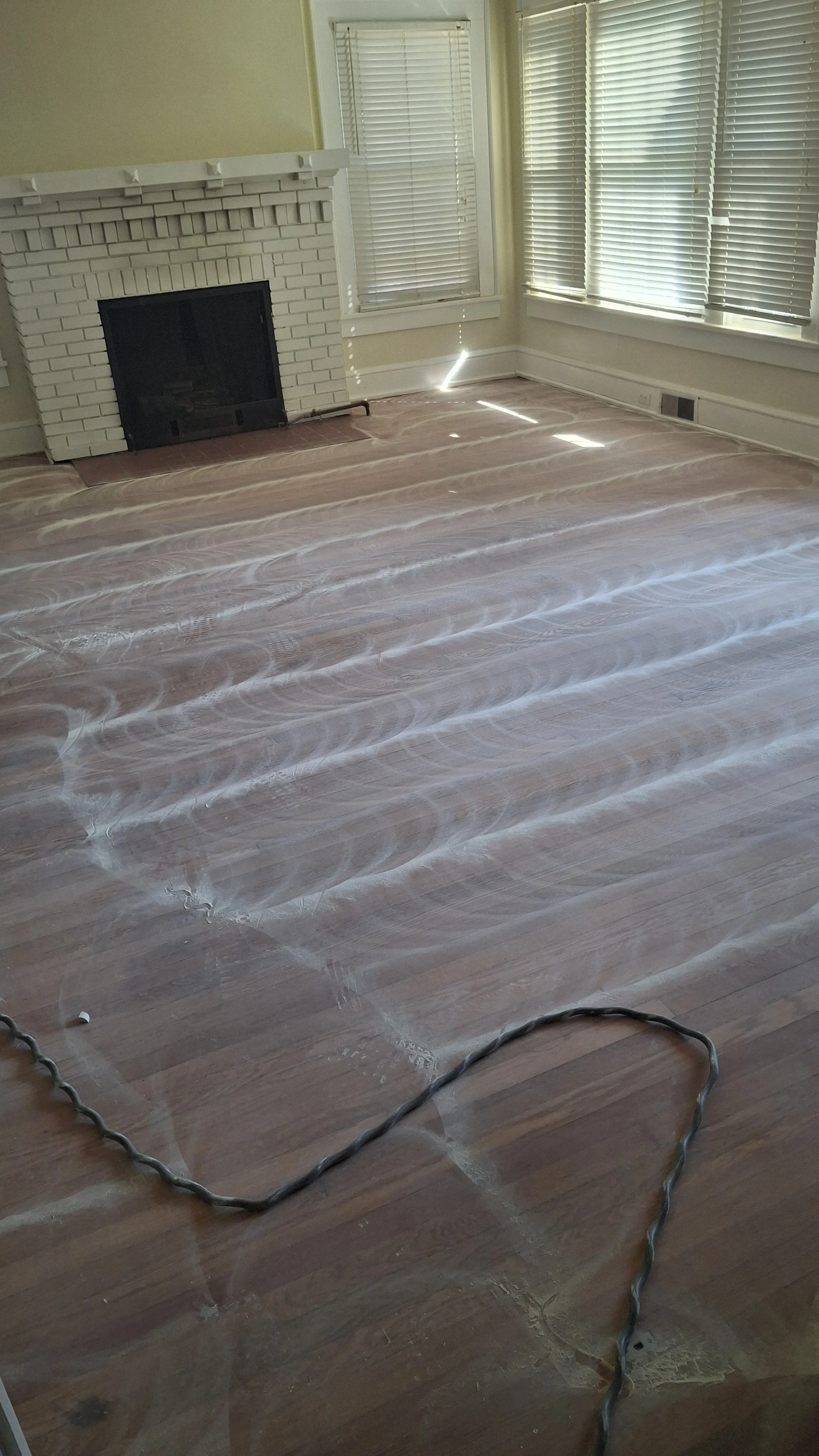 Empty room with hardwood flooring, a white brick fireplace, and window blinds. The floor has dried grout for tile installation. Power cords are on the floor.