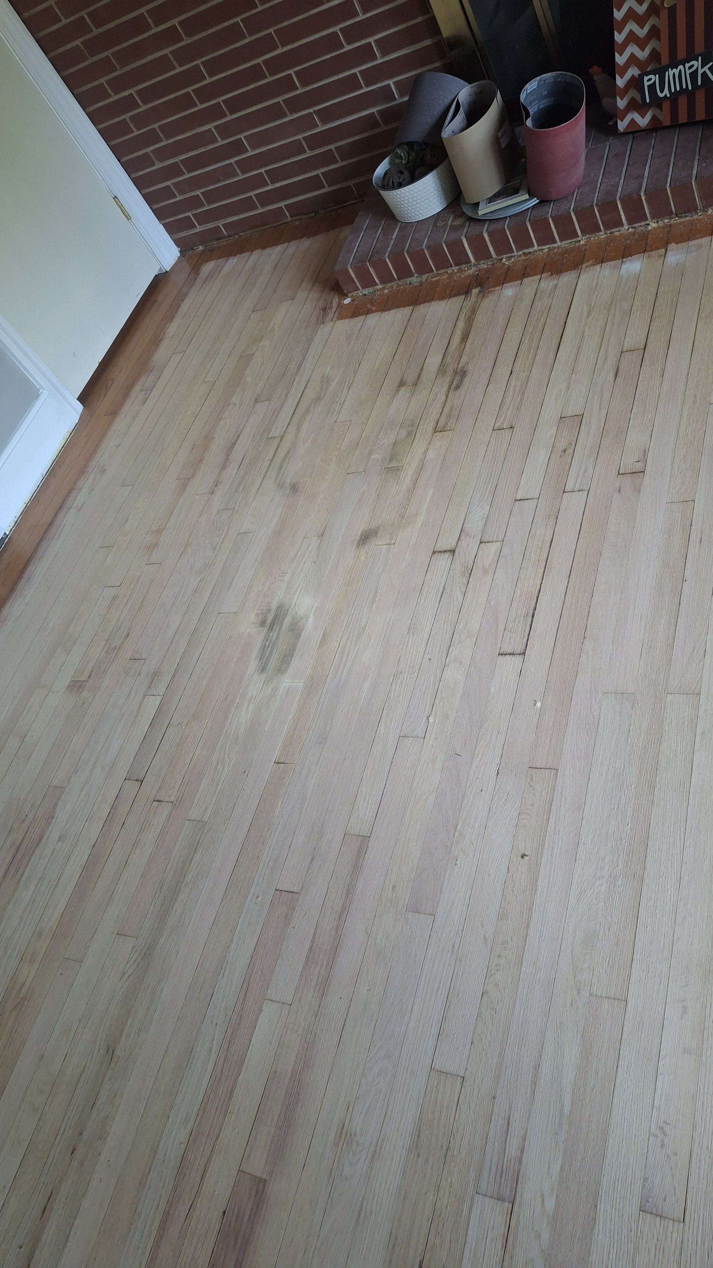 Photo of a light-colored wooden floor with some dirt marks, near a brick fireplace with a small brick step. Items on the brick step include rolled-up posters or papers, a book, and a plant. A sign with the word 'PUMPKIN' is partly visible in the back