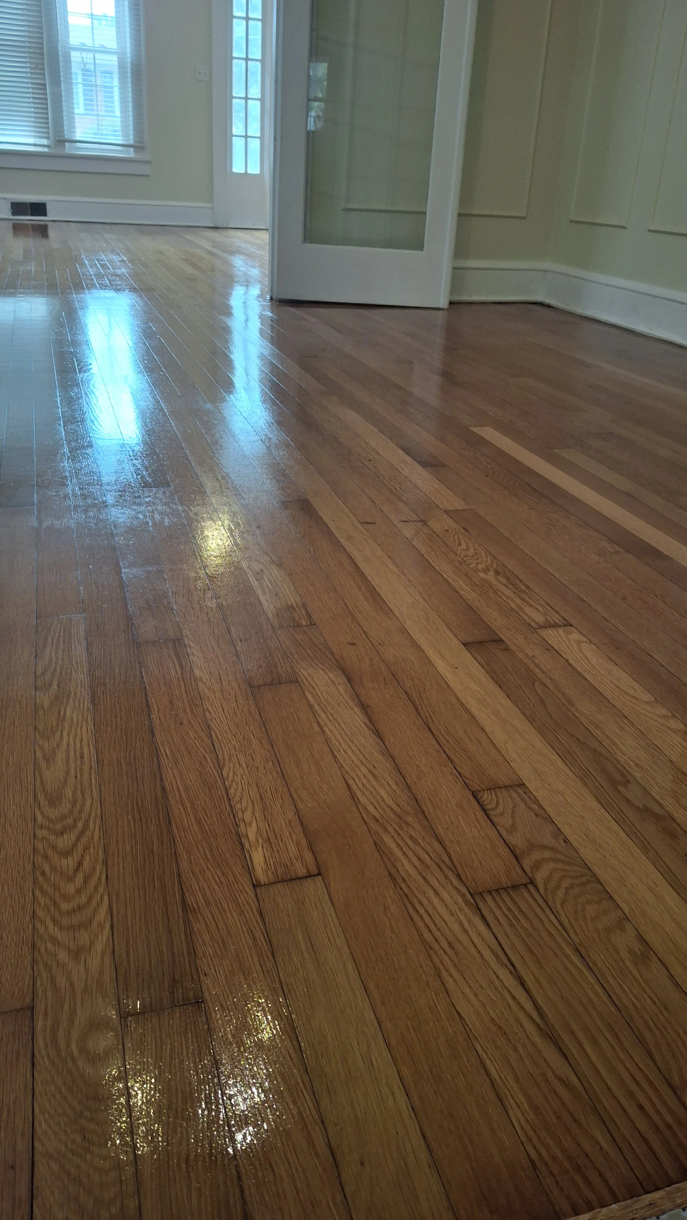 A room with shiny, polished hardwood floors, cream-colored walls, a closed French door, and a window with blinds.