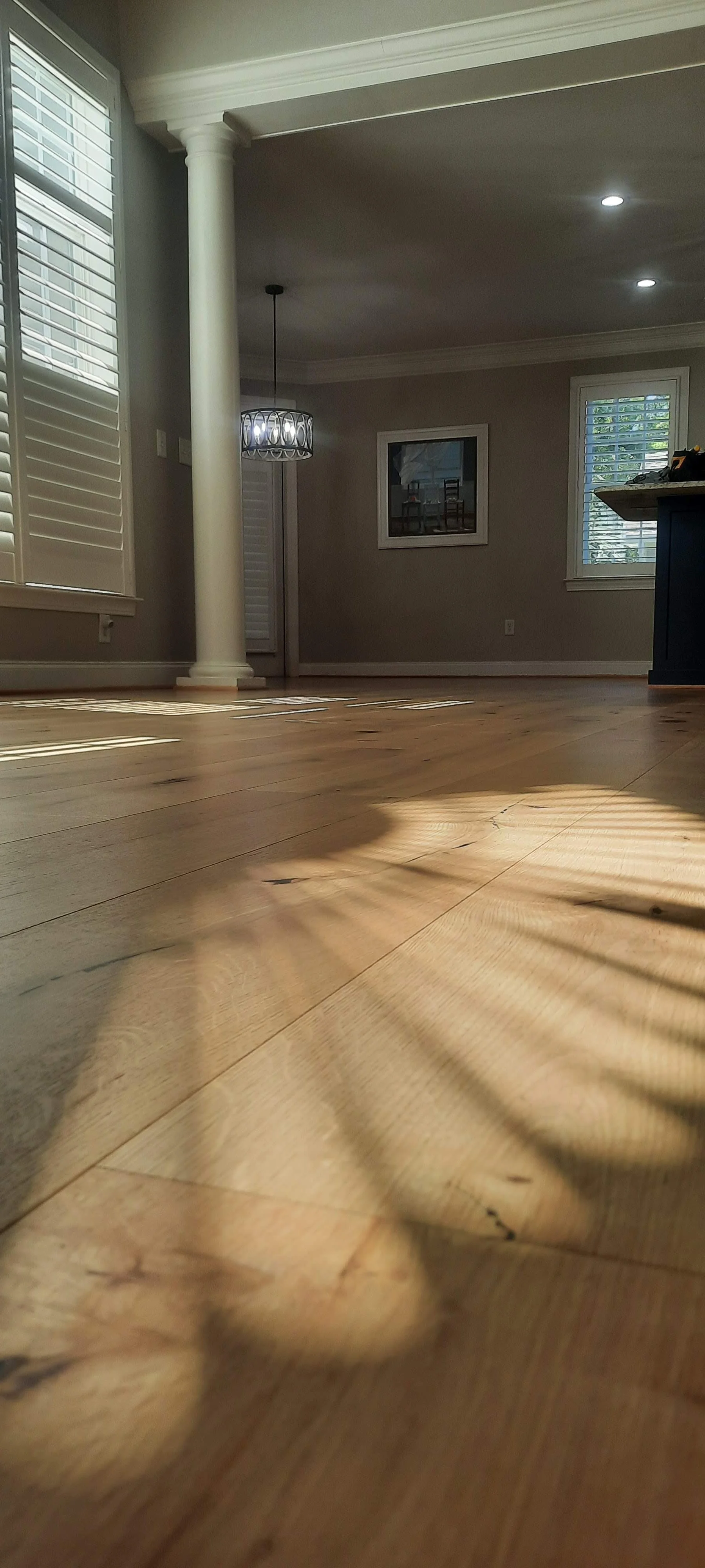 Empty living room with sunlight streaming through window shutters, casting shadows on a light hardwood floor, promoting Floyd's Flooring & Stairs LLC