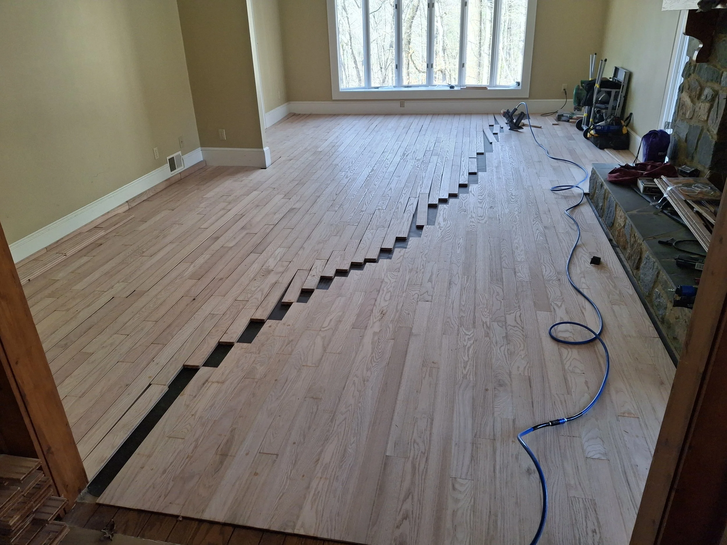A room with large windows, yellow walls, and hardwood flooring in the process of being refinished, with some floorboards lifted and construction tools scattered around.