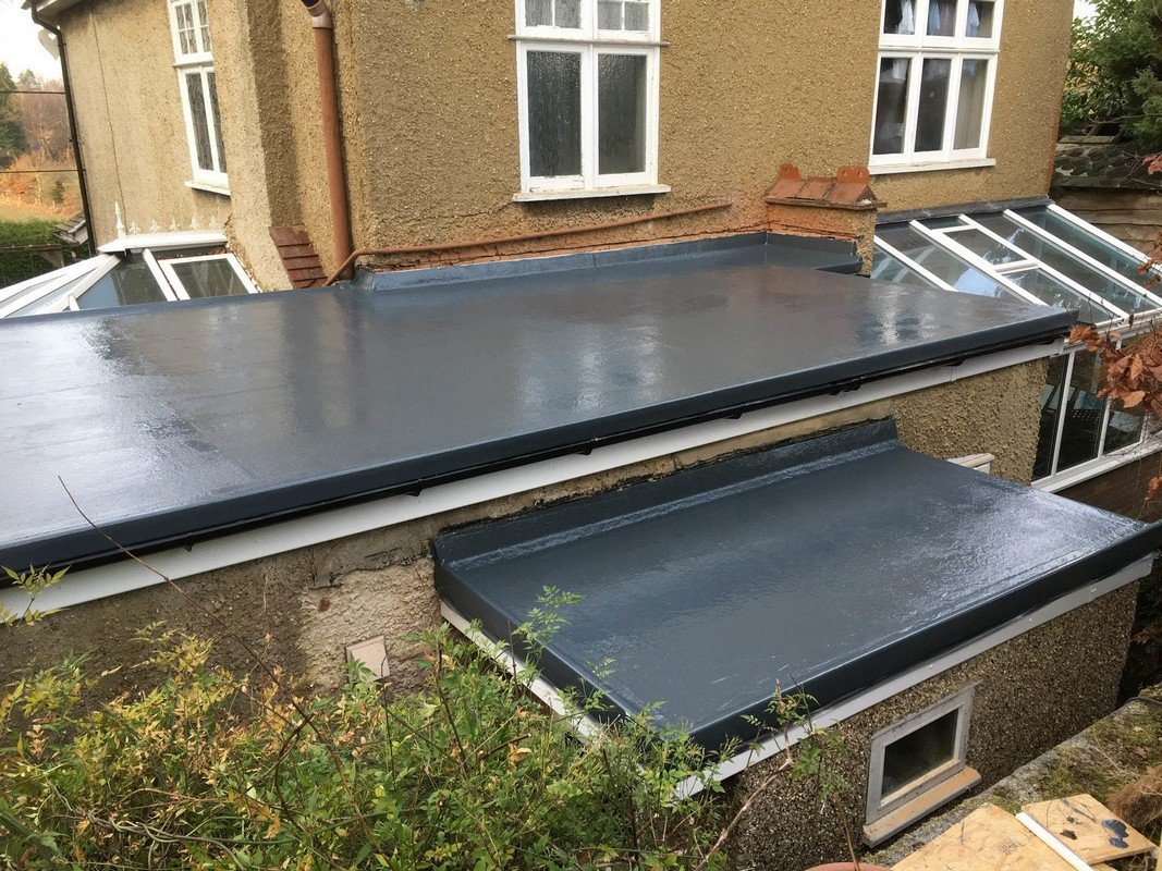 The image shows the flat roof of a building with new dark-colored waterproof coating, surrounded by multiple windows and some plants in the foreground.
