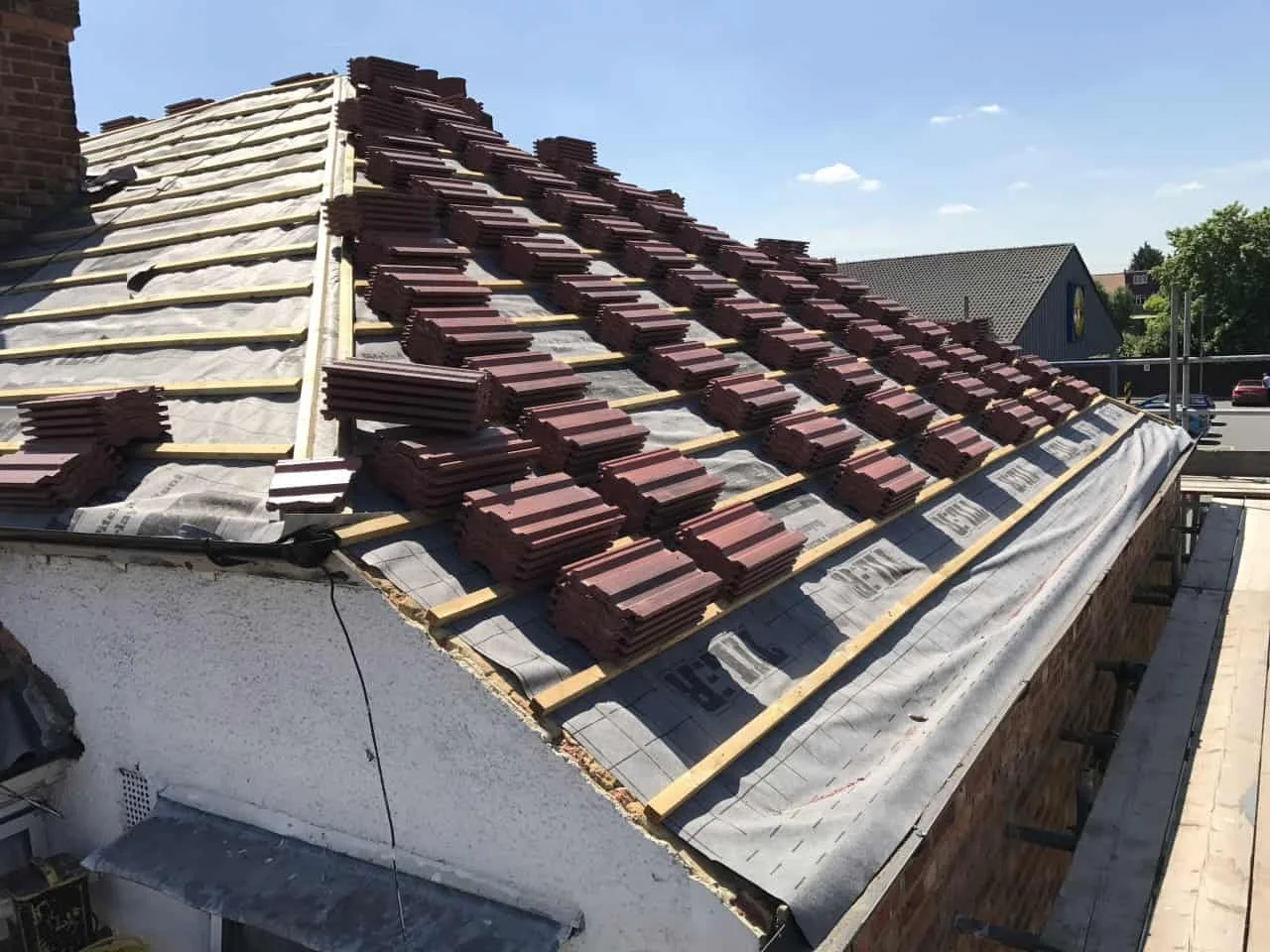 Roof under construction with stacks of red roof tiles and wooden battens installed, partially covered with protective sheeting.