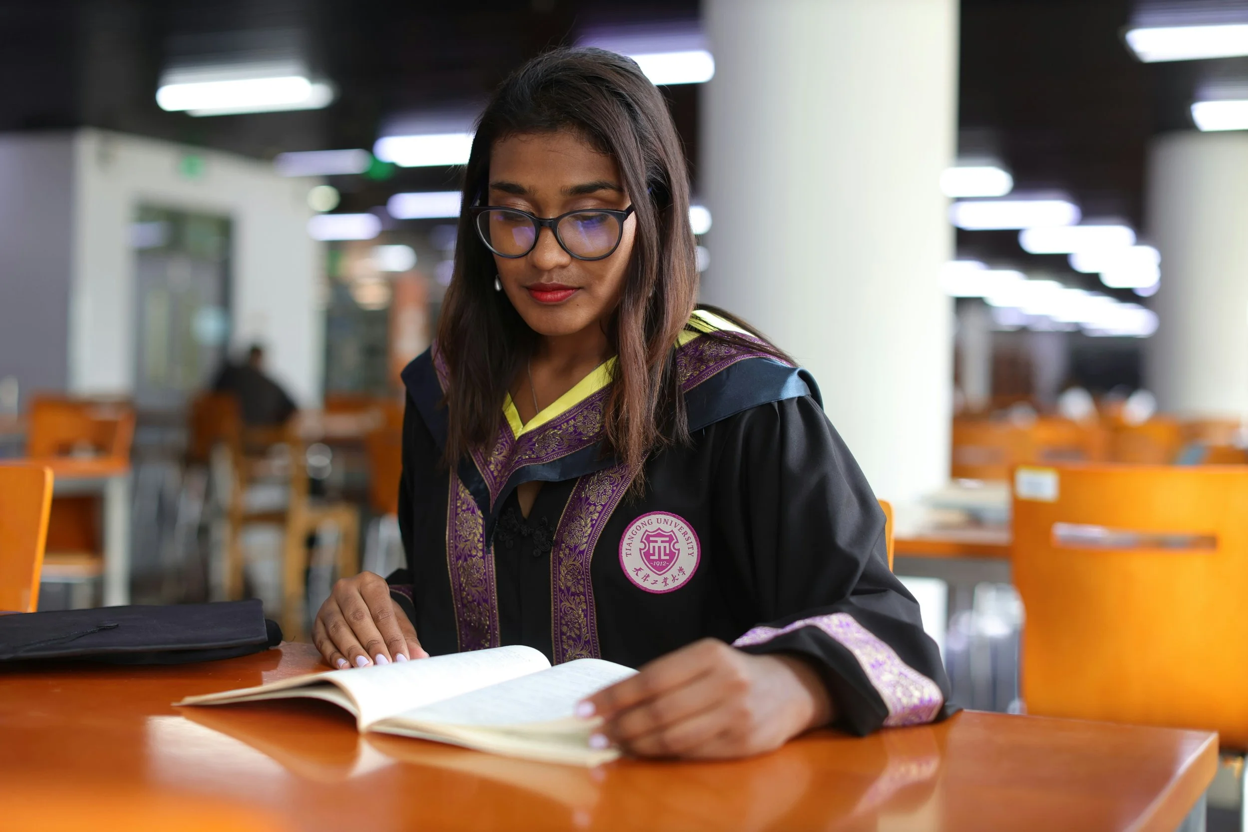 A woman wearing glasses and a graduation gown with an emblem from Tiangong University on it, reading a book at a desk inside a library or study area with orange chairs and bright overhead lighting.
