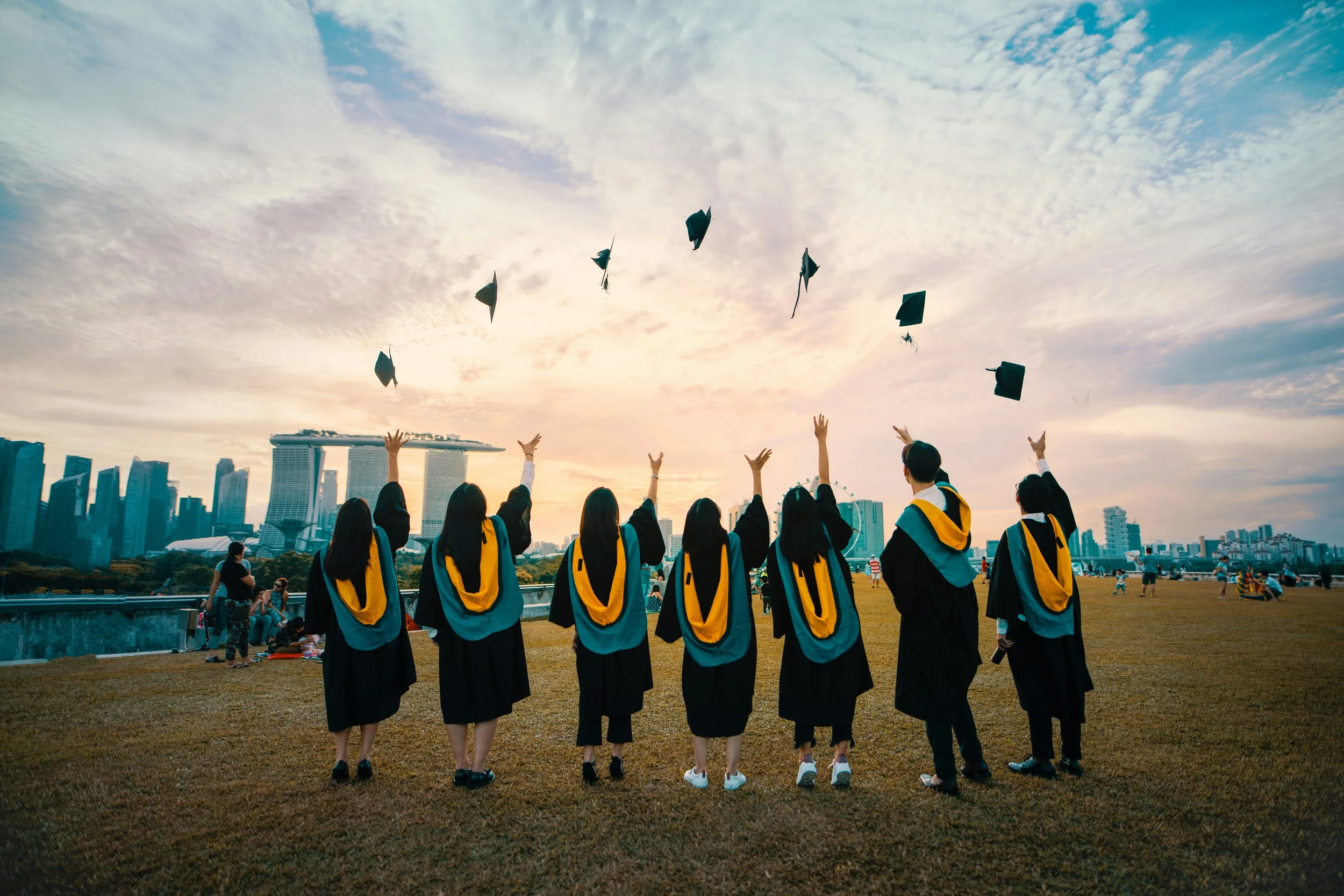 Group of graduates in black and yellow robes tossing caps into the air during a graduation ceremony outdoors with cityscape in the background at sunset.