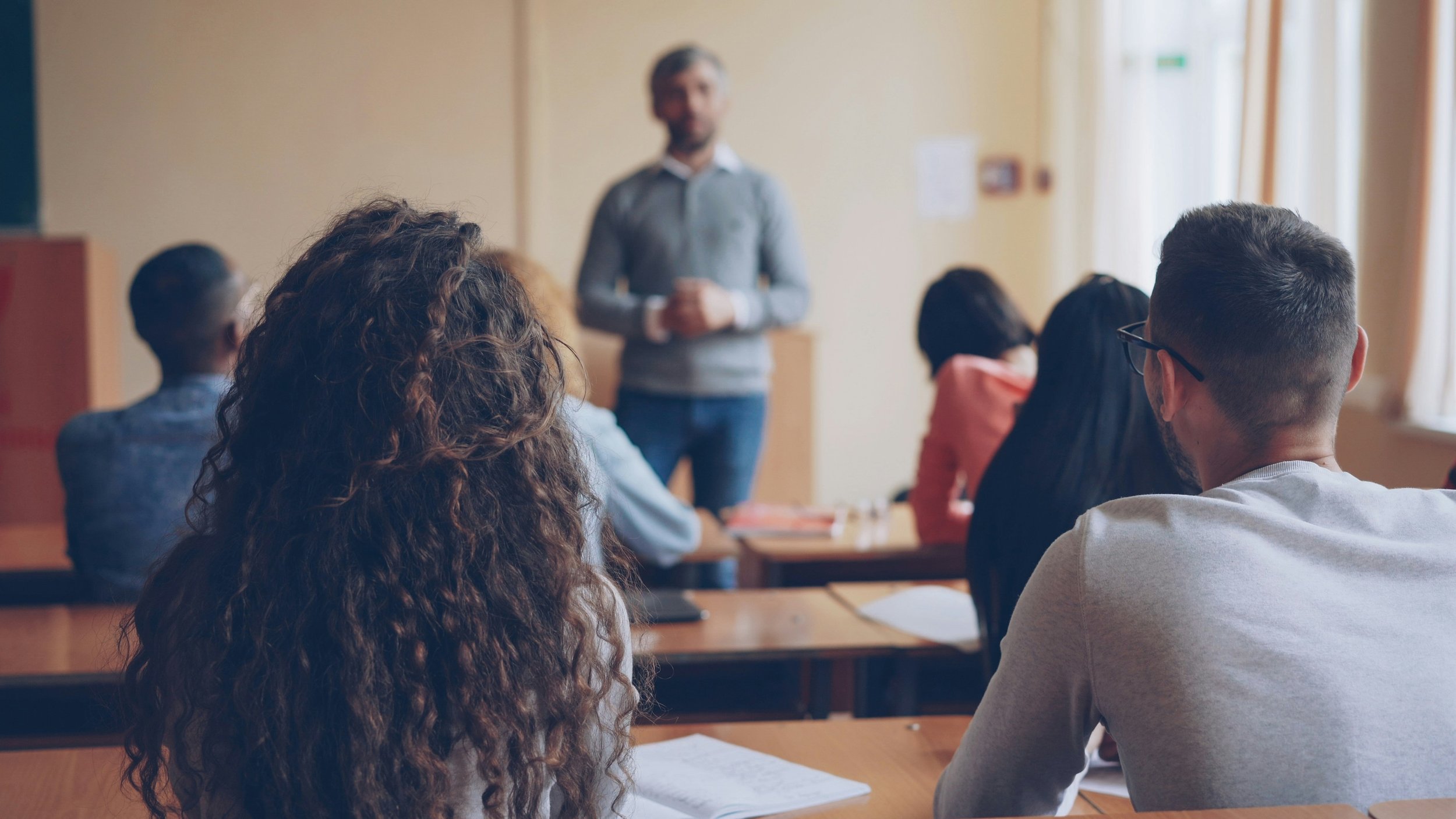 A classroom with a male teacher standing at the front, and students seated facing him, some taking notes or listening.