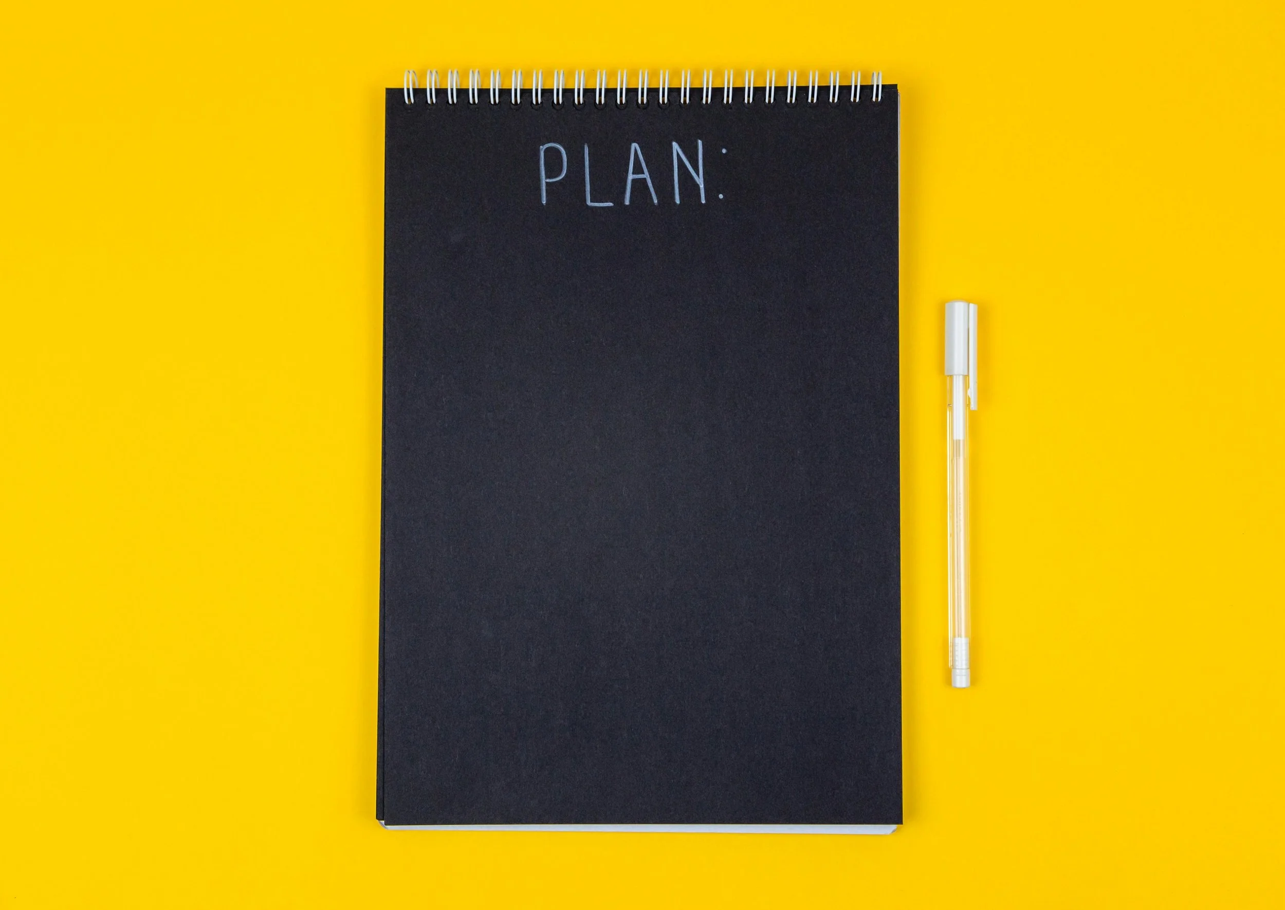 A black spiral-bound notebook with the word "PLAN" written on the cover, accompanied by a white marker pen, set against a bright yellow background.