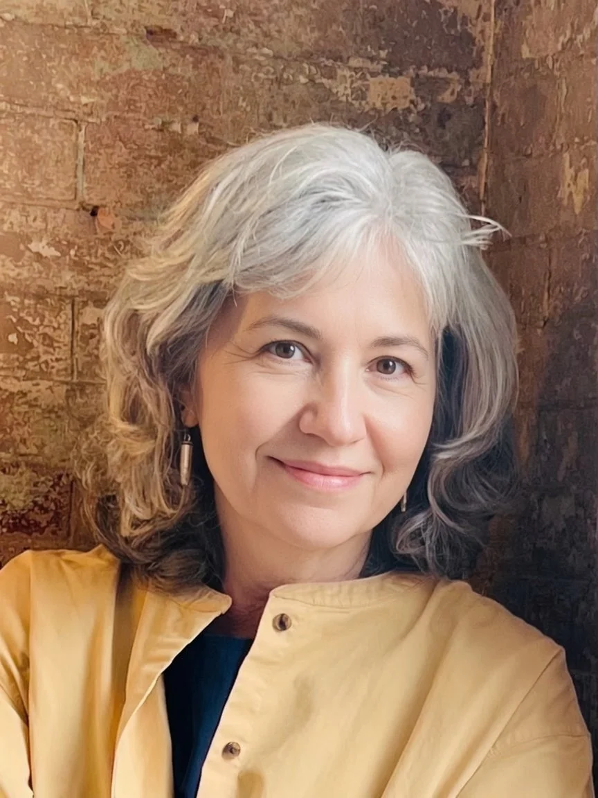 A woman with shoulder-length gray hair smiling at the camera, wearing a yellow jacket and earrings, sitting in front of a brick wall.