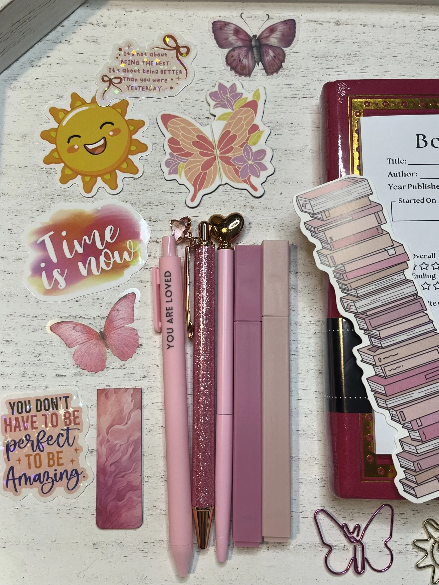 Pink and glittery pens, decorative stickers including a smiling sun, butterfly, and motivational quotes, along with a red book with a sticker of stacked books, all arranged on a white wooden surface.
