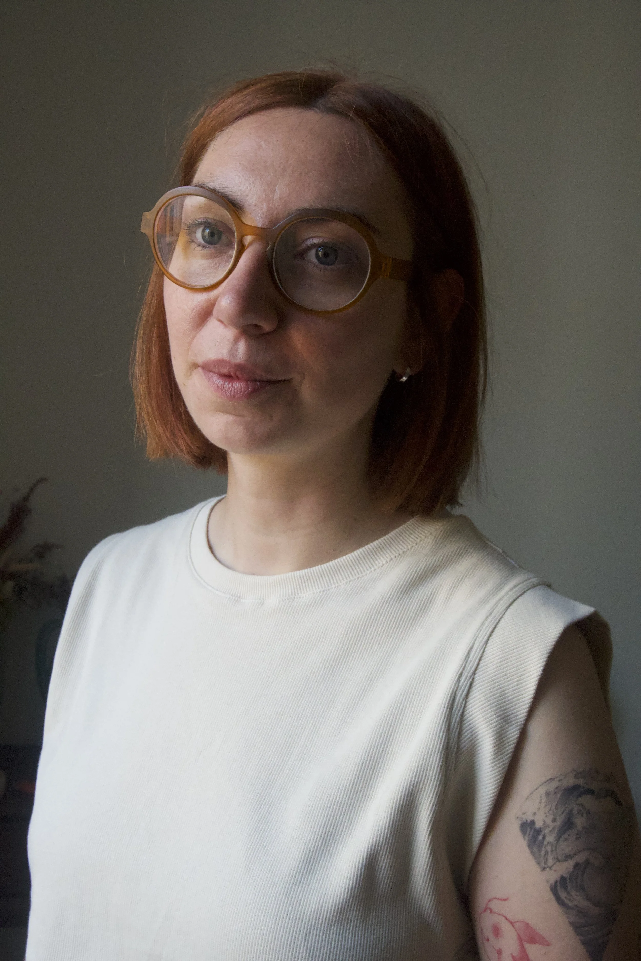 Une femme avec des lunettes rondes translucides et une t-shirt blanc, montrant son bras tatoué, dans un intérieur