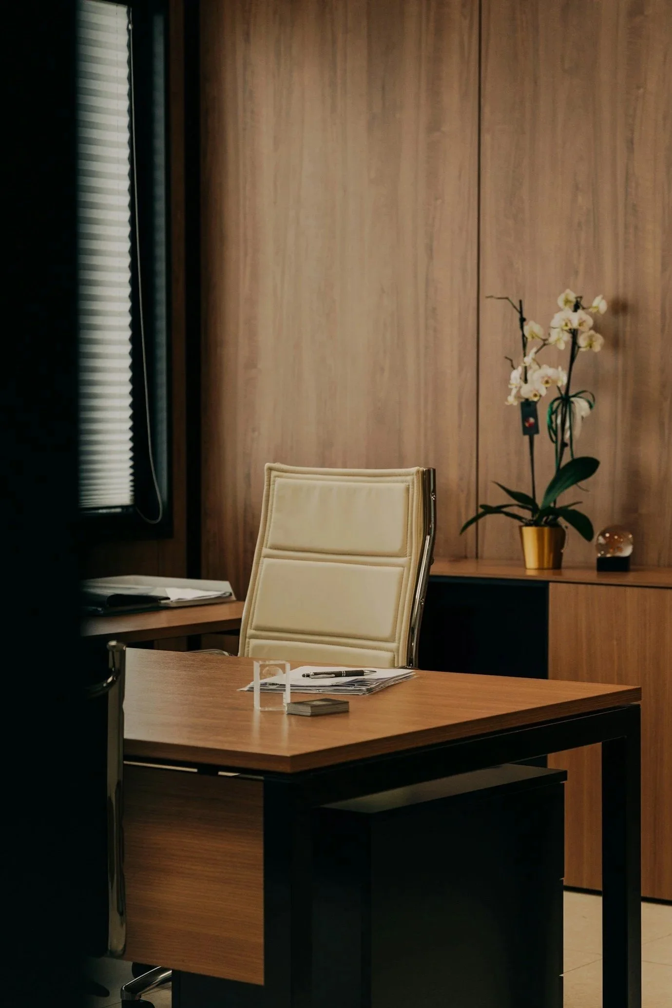 Un bureau avec une chaise blanche, une table en bois avec des papiers, un stylo, et une décoration avec une plante orchidée dans un pot doré, en arrière-plan un mur en bois.