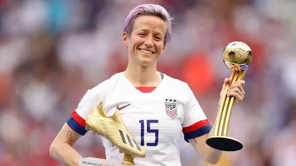 Megan Rapinoe, legendary openly lesbian professional soccer player, smiling and holding the FIFA Women's World Cup trophy in her USA jersey.