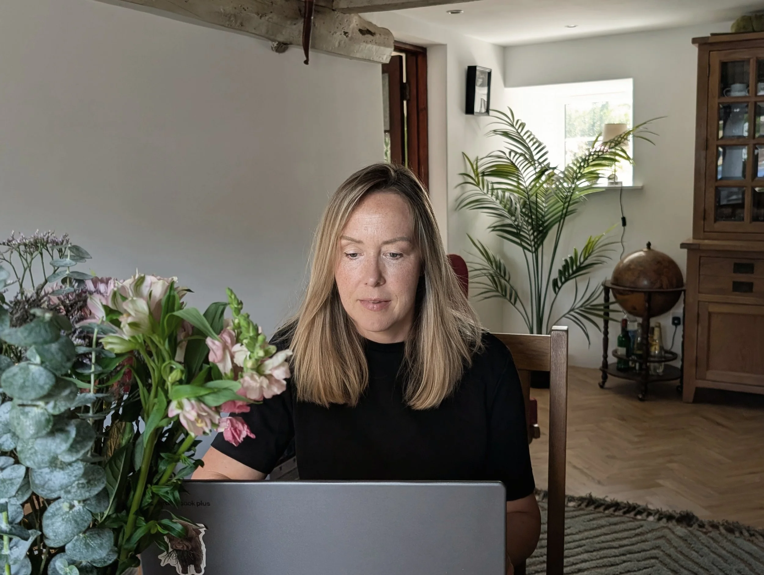 A woman with long blonde hair sitting at a dining table, working on a laptop, surrounded by a bouquet of pink and white flowers, in a bright room with a large window, a potted plant, and wooden furniture in the background.