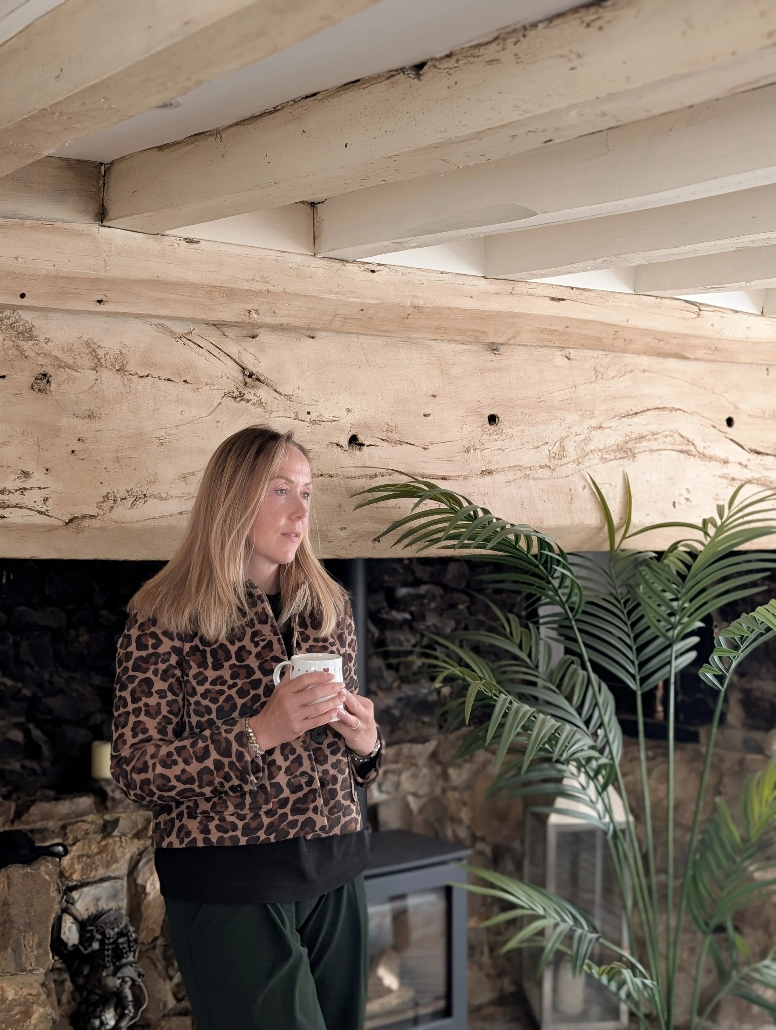 A woman with long blond hair standing indoors, holding a white mug, wearing a leopard print jacket and dark pants, near a large green potted plant, with a wooden wall and stone fireplace behind her.