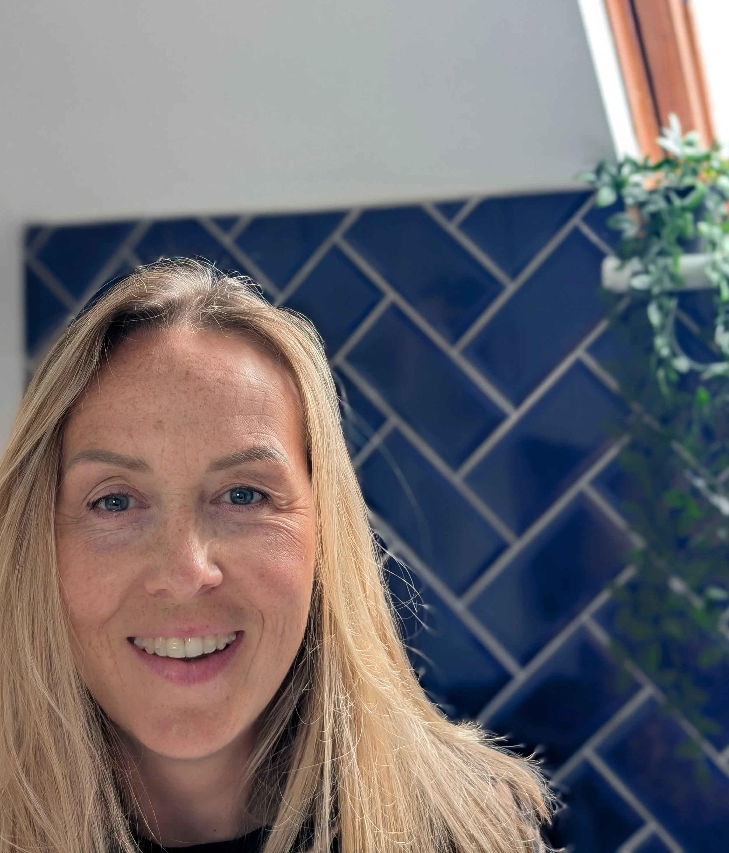 A woman with blonde hair smiling at the camera. Part of her face, including eyes, nose, mouth, and hair, is visible. The background features a blue tiled wall and a window with a plant on the right side.