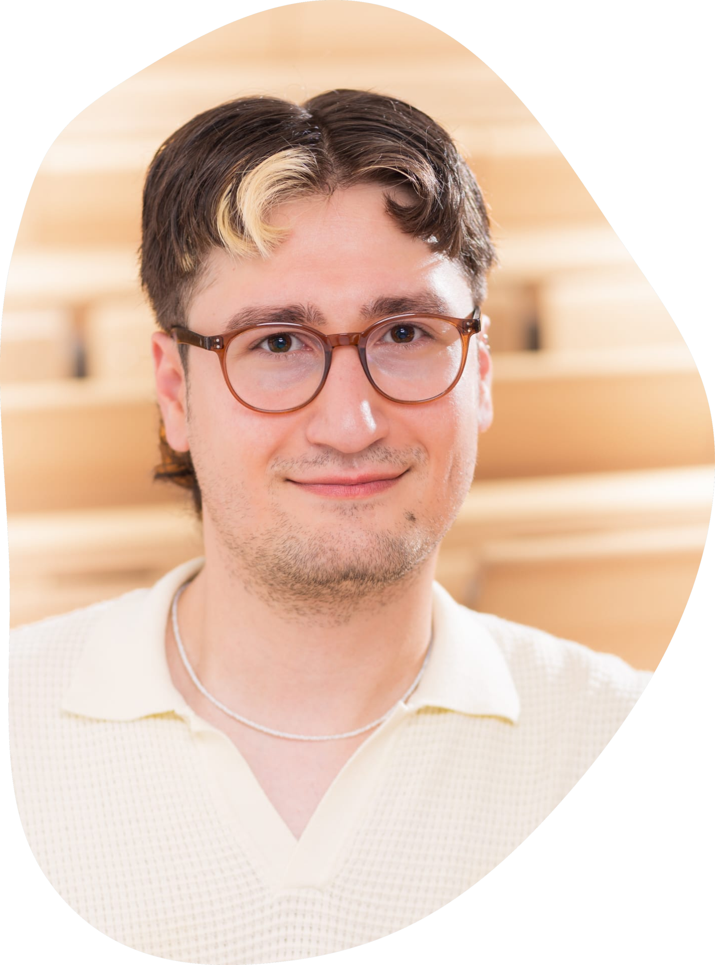 Close-up of a young man with brown and blonde hair, wearing brown glasses and a yellow polo shirt, smiling softly in front of a wooden background.