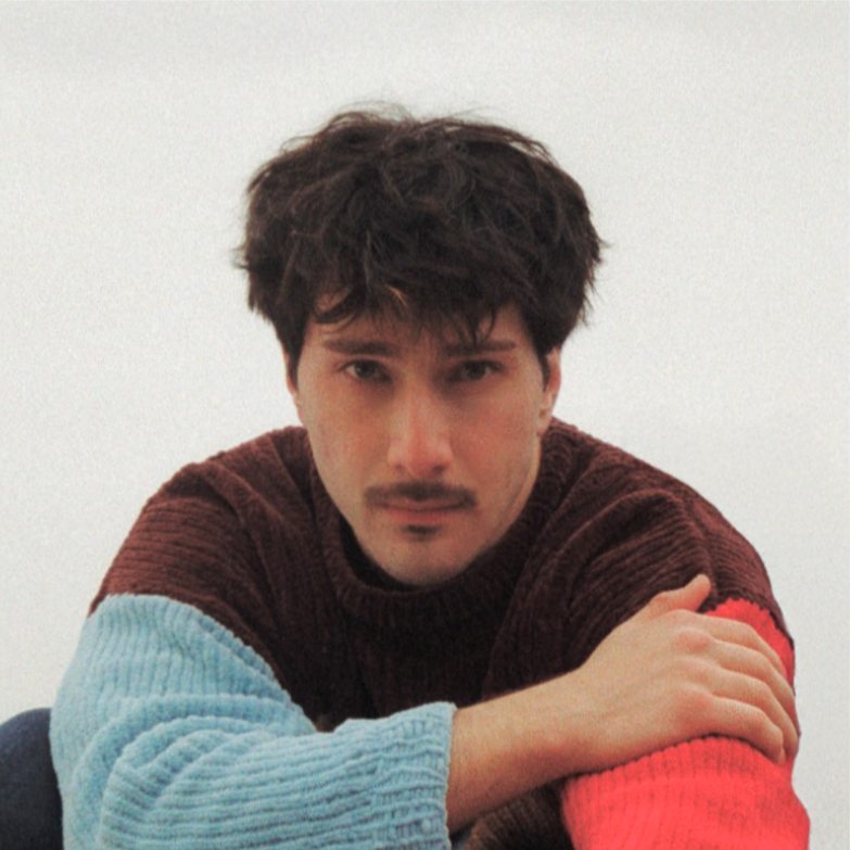 A young man with dark, messy hair and a mustache wearing a multicolored sweater, sitting against a plain white background.
