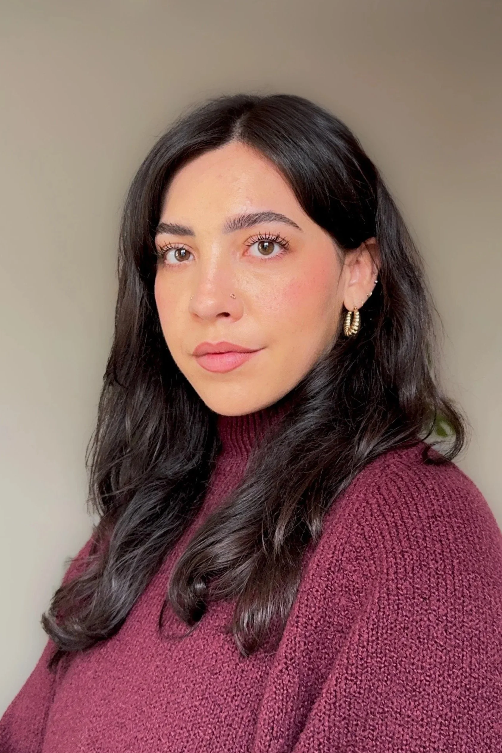 A woman with long dark hair, wearing hoop earrings and a maroon sweater, looking at the camera with a neutral expression against a plain beige wall.