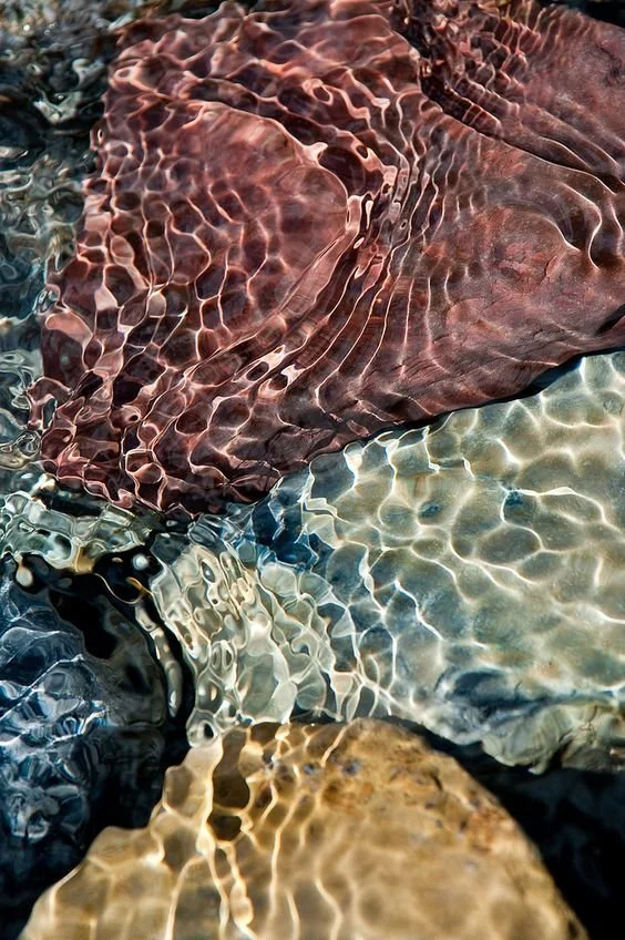 Close-up of colorful rocks submerged in clear water, with sunlight creating shimmering reflections on the surface.