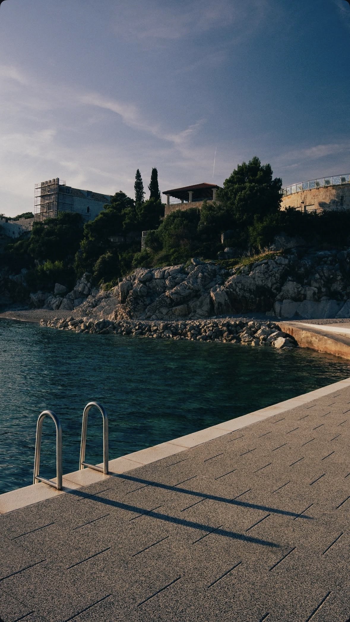 A seaside view with a swimming pool in the foreground, rocky shoreline, and houses with trees on a hill in the background under a partly cloudy sky.