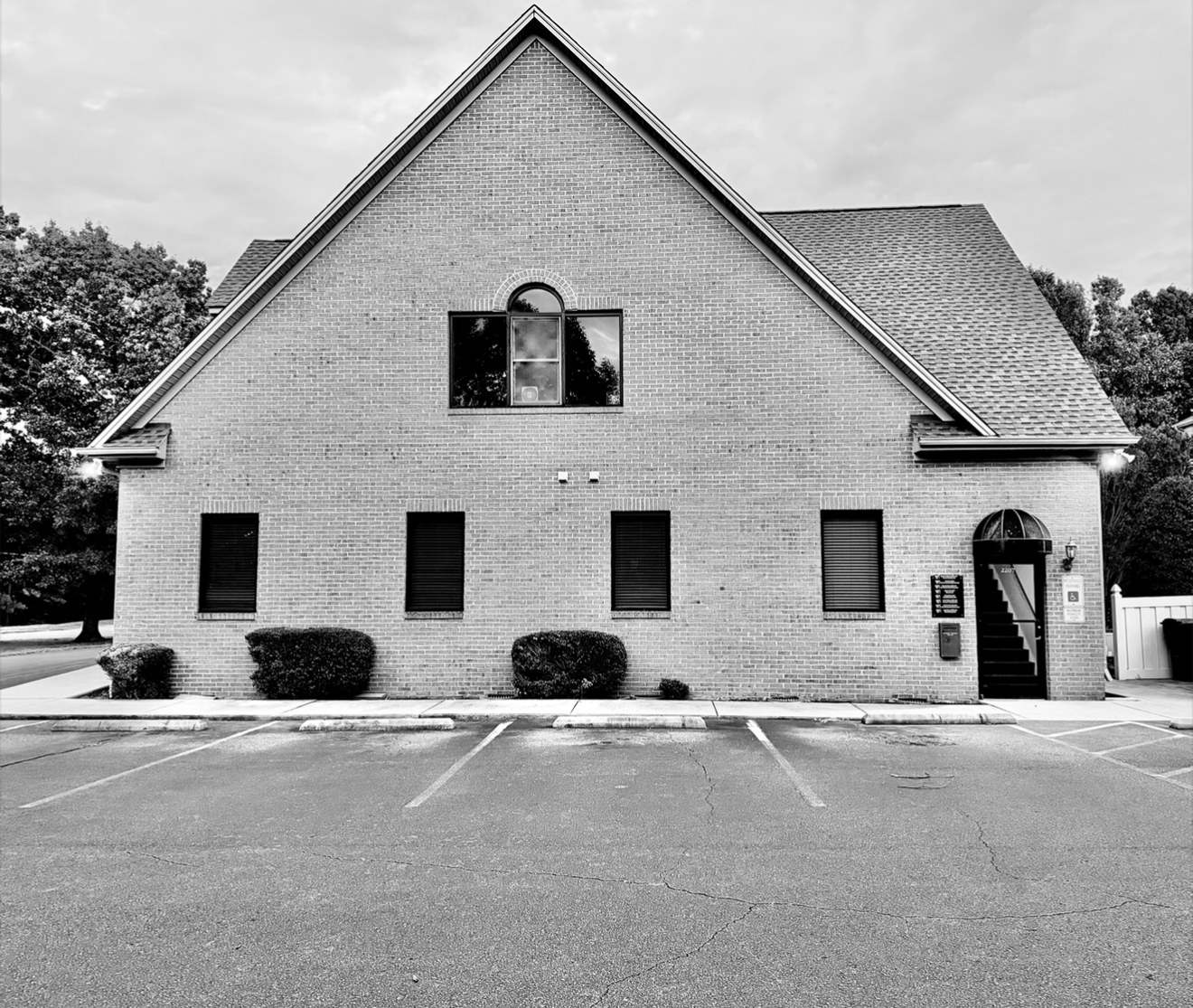 Black and white photo of a brick building with a steep roof, four small windows on the ground floor, one large arched window on the upper floor, and door leading to the entrance  of Sxuations PLLC. Parking lot in front.
