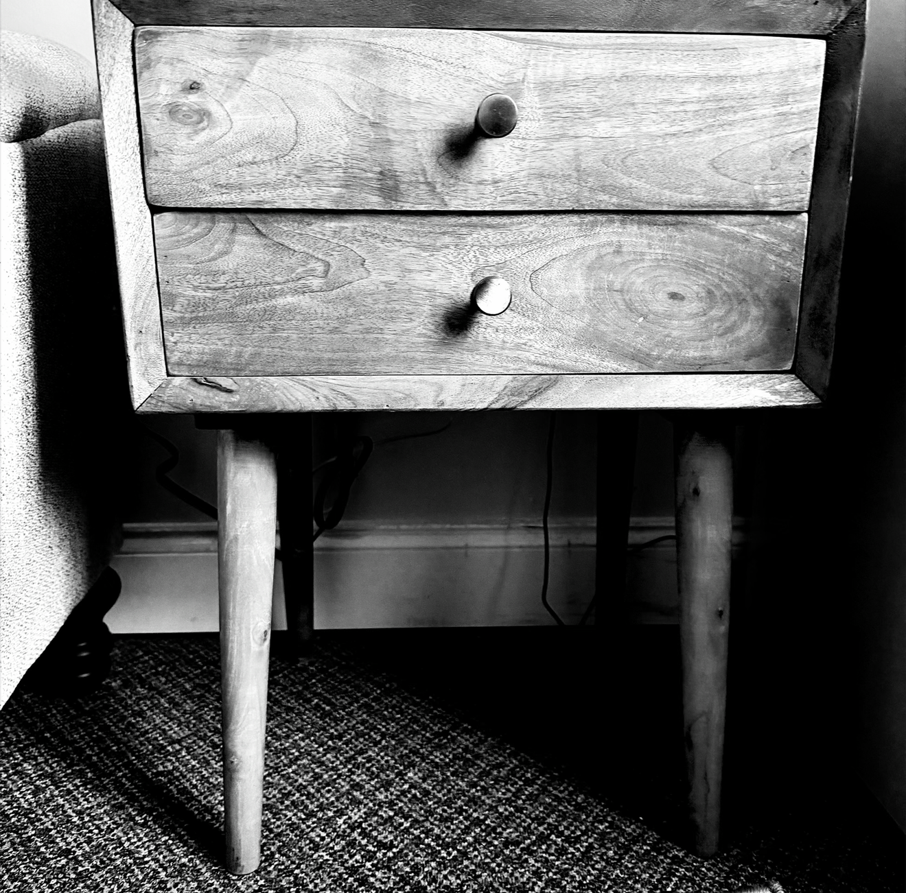 Black and white photo of a wooden bedside table in therapy office Sxuations PLLC with two drawers and round knobs, standing on angled legs on a textured carpeted floor, against a wall with a power outlet and some cords visible underneath.
