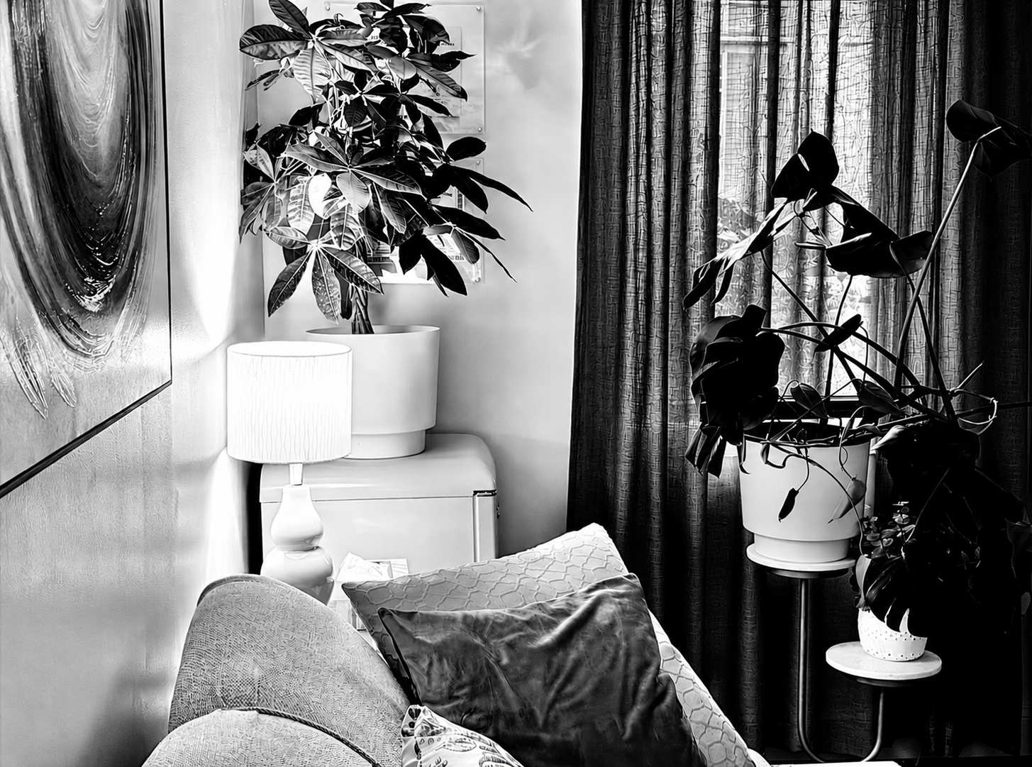 A black-and-white photo of the interior of the therapy practice Sxuations, includes a sofa, cushions, a lamp, houseplants in pots, curtains, and  wall art. Photo by Taneka M. Bennett for Sxuations.