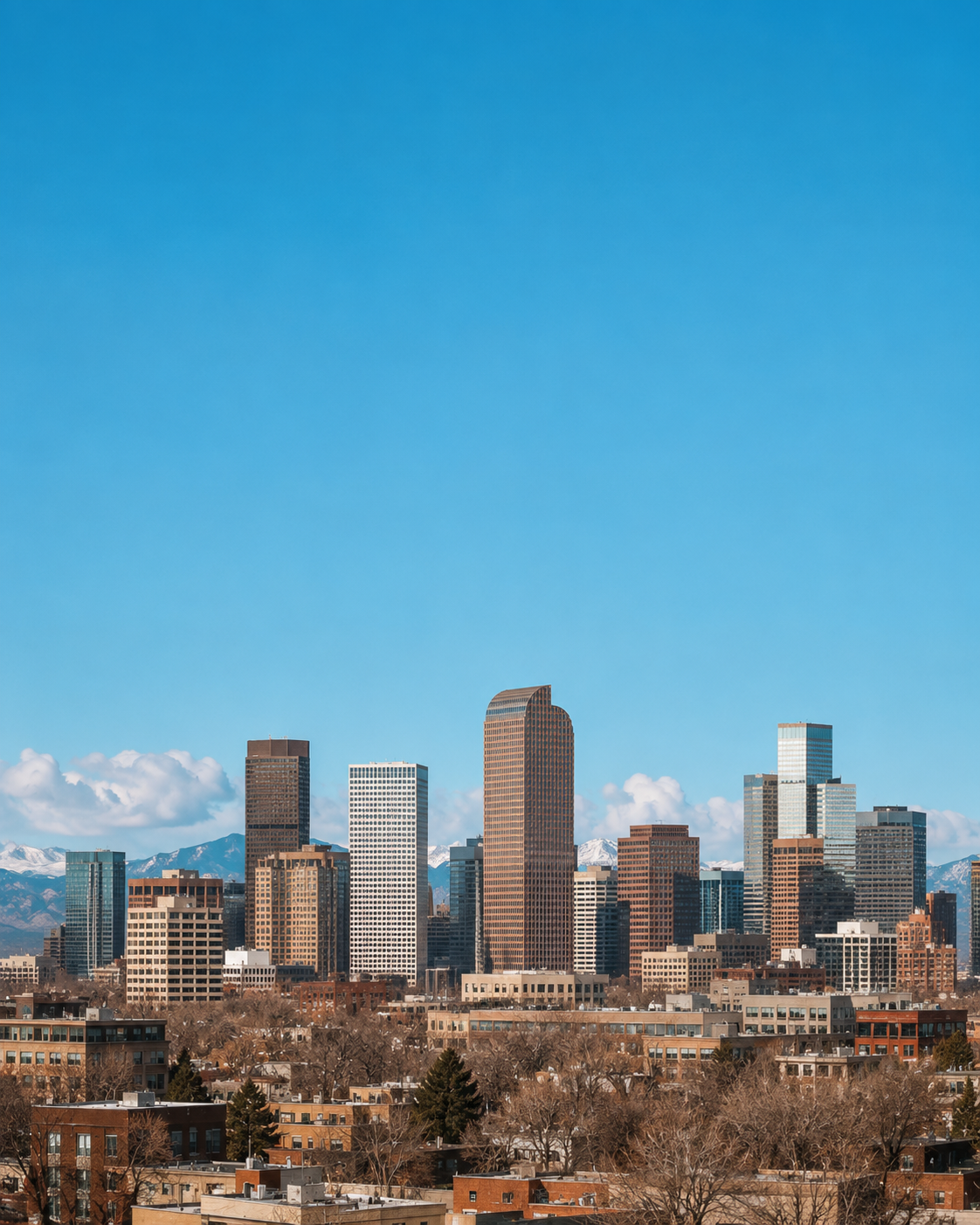 City skyline with tall skyscrapers against a bright blue sky and snow-capped mountains in the background, with trees and residential buildings in the foreground.