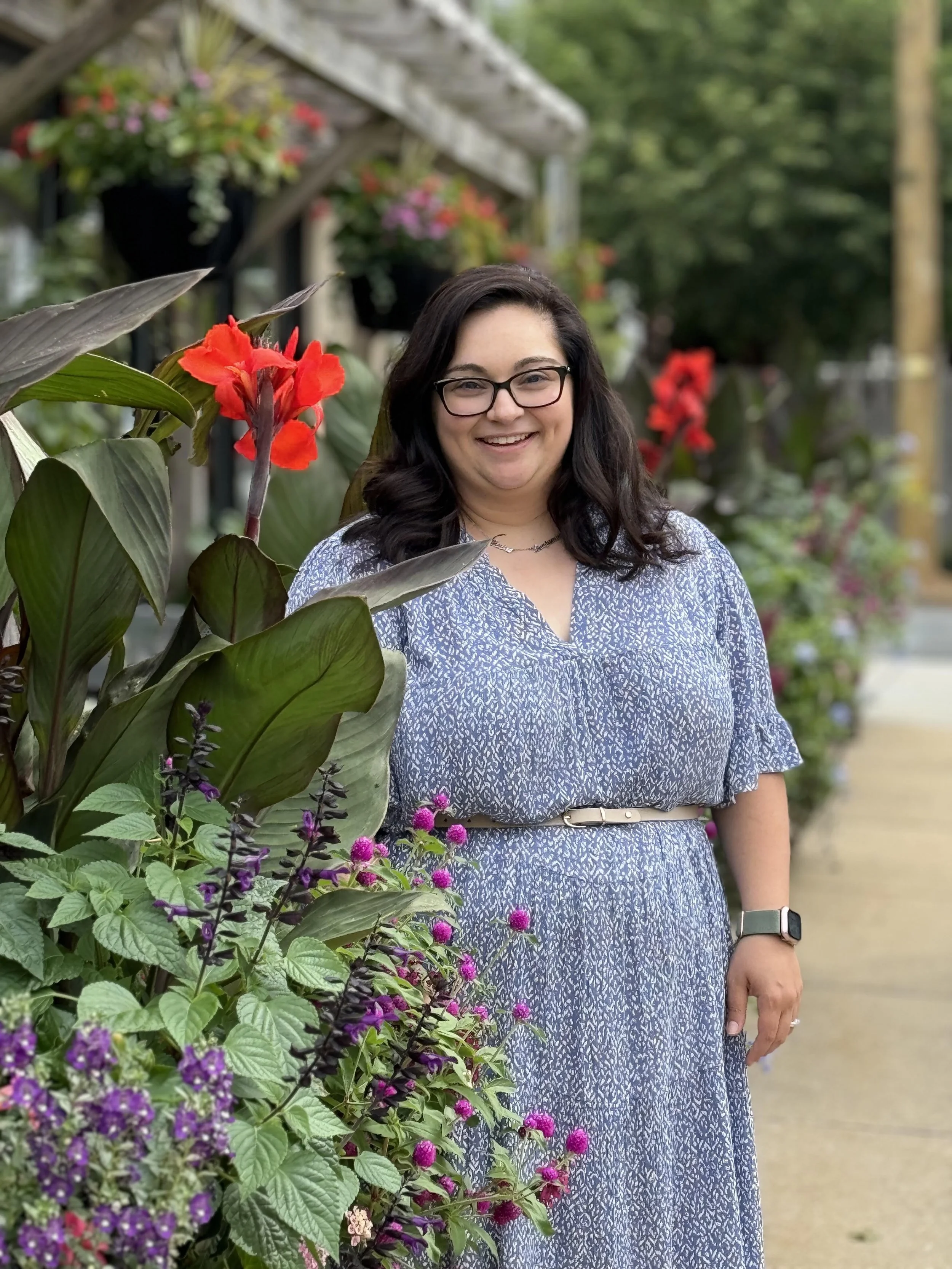 A smiling woman with dark hair and glasses standing outdoors next to colorful flowers, including red and purple blossoms, on a sunny day with a blurred background of greenery and a sidewalk.