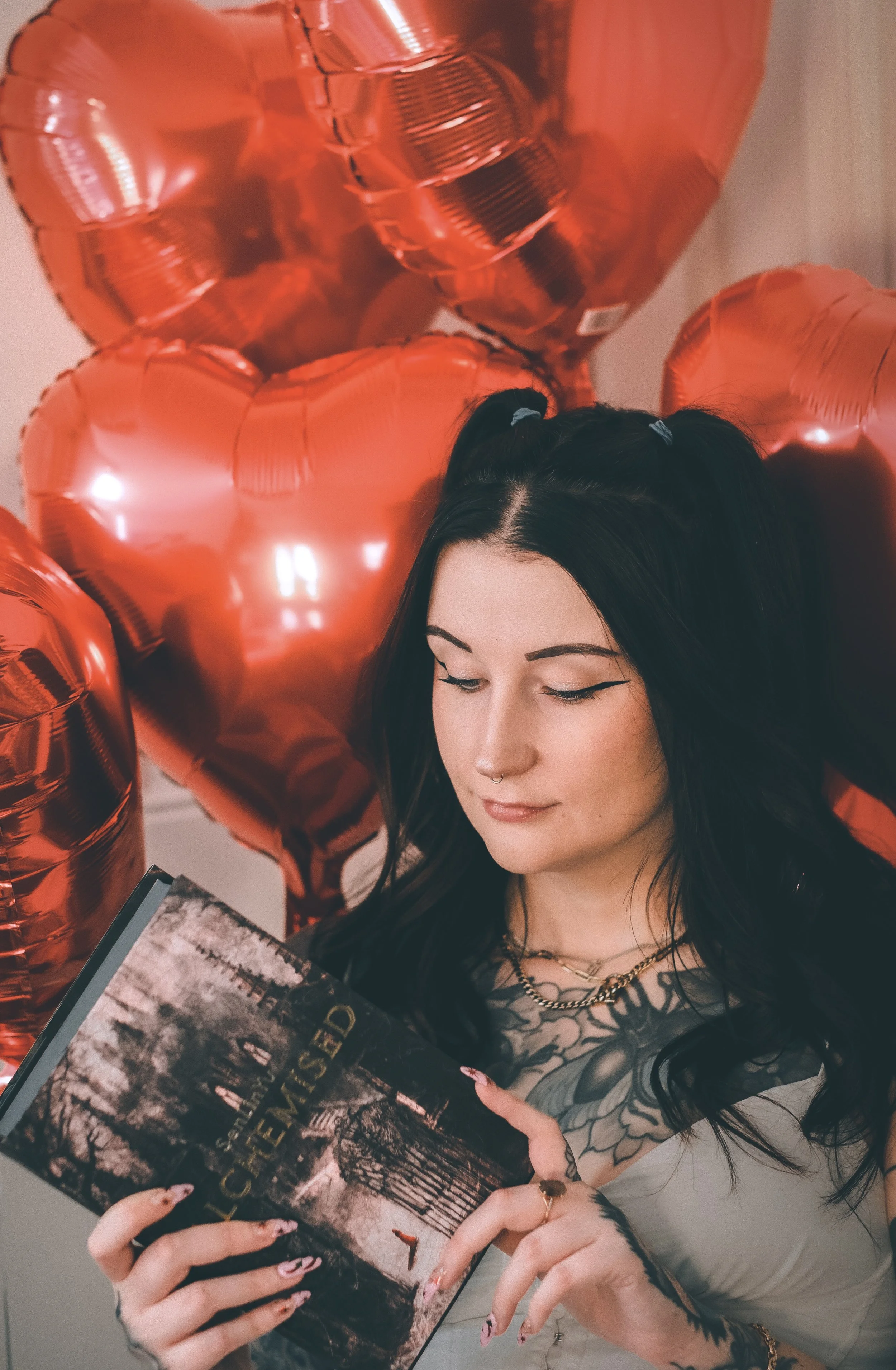 A young woman with black hair styled in pigtails, wearing winged eyeliner, a septum piercing, multiple necklaces, and rings, is sitting at a table reading a photo book titled "Alchemised" She is surrounded by shiny red heart-shaped balloons.