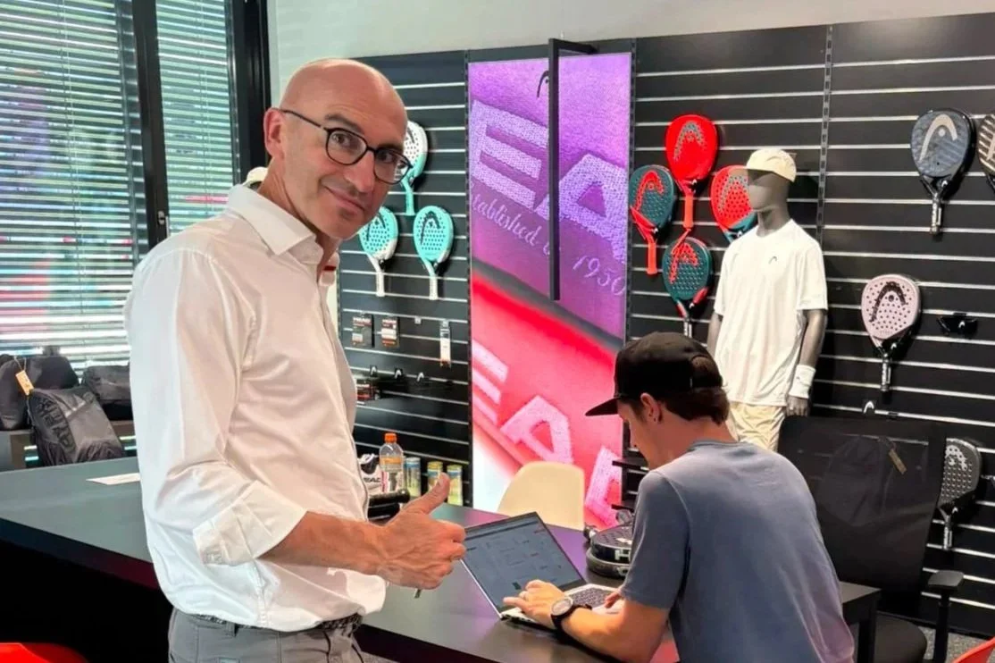Ein Mann mit Brille und weißen Hemd steht in einem Laden, der Tennis- und Badmintonschläger verkauft, und zeigt auf die Kamera, während eine Person am Laptop arbeitet.