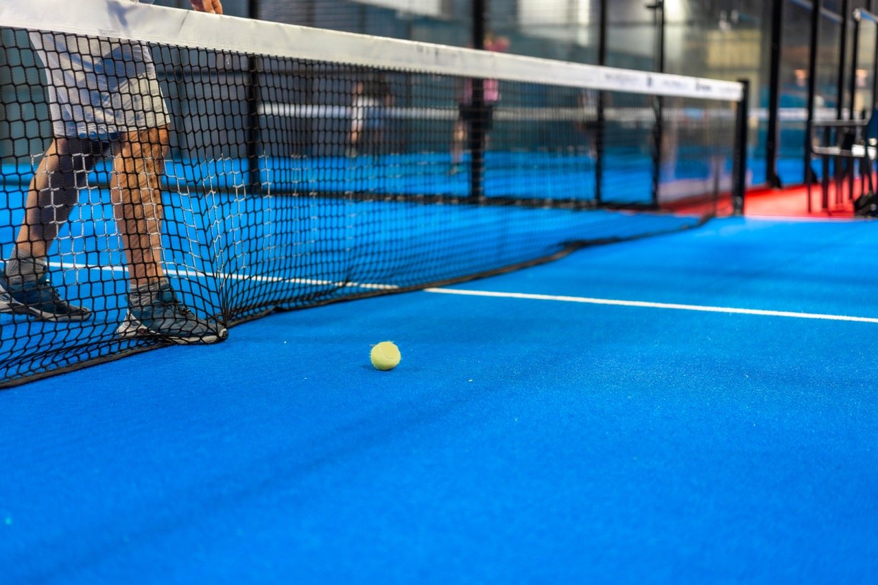 Boden eines Tennisplatzes in Blau mit Tennisball und Netz, im Hintergrund sind Menschen.