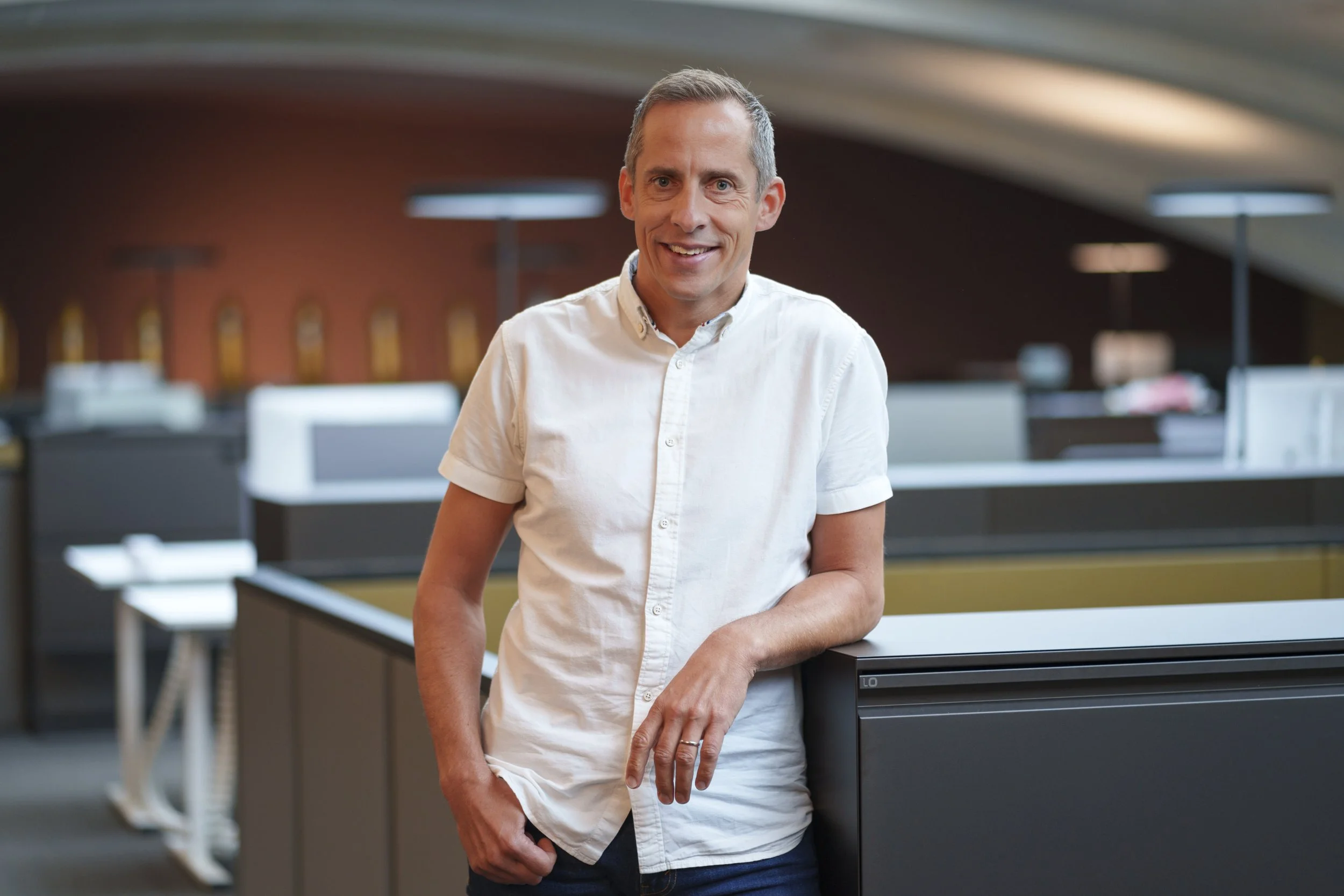 Ein lächelnder Mann in einem weißen Hemd steht in einem Büro mit modernen Büroarbeitsplätzen im Hintergrund.