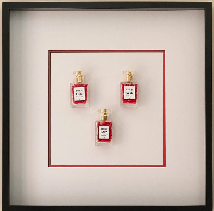 Three small perfume bottles labeled "Dose of Love" are arranged in a triangle on a white display with a red border, inside a black frame.